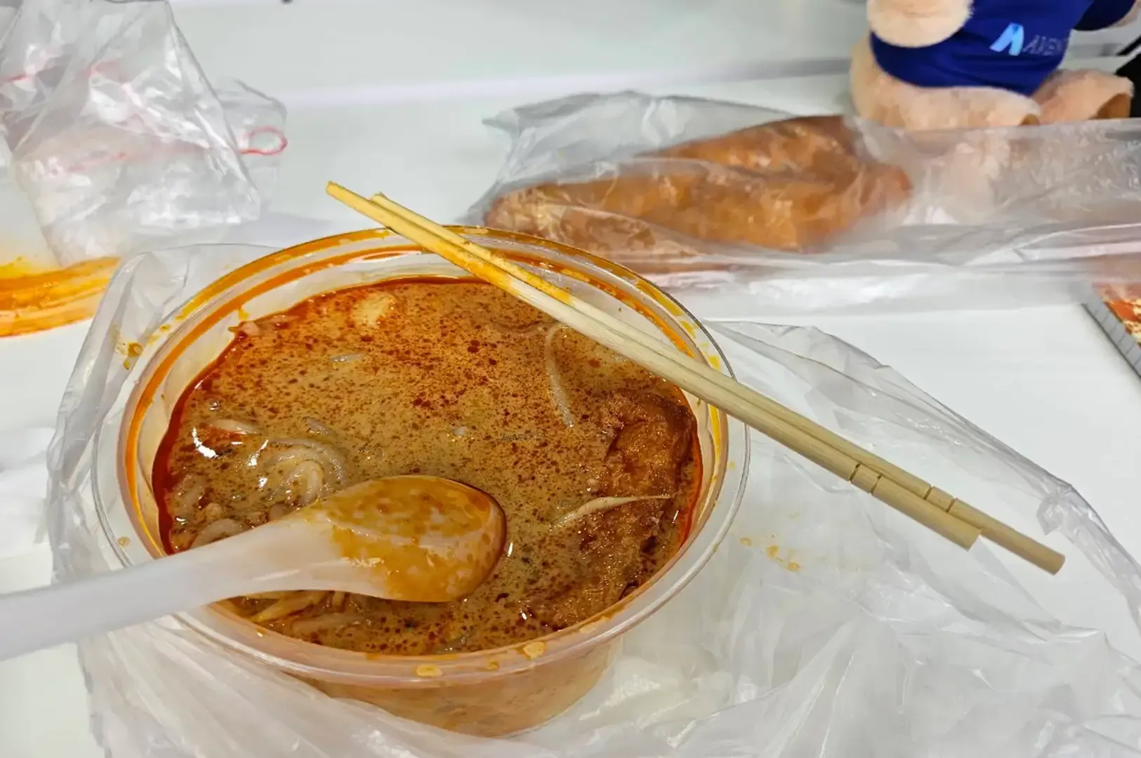 Takeout bowl of spicy soup with spoon and chopsticks on a plastic-covered table