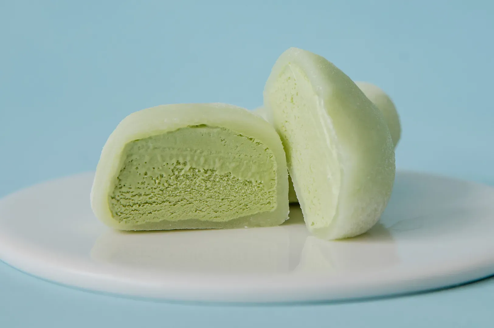 Two pale green steamed buns on a white plate, one cut open to show a soft filling.