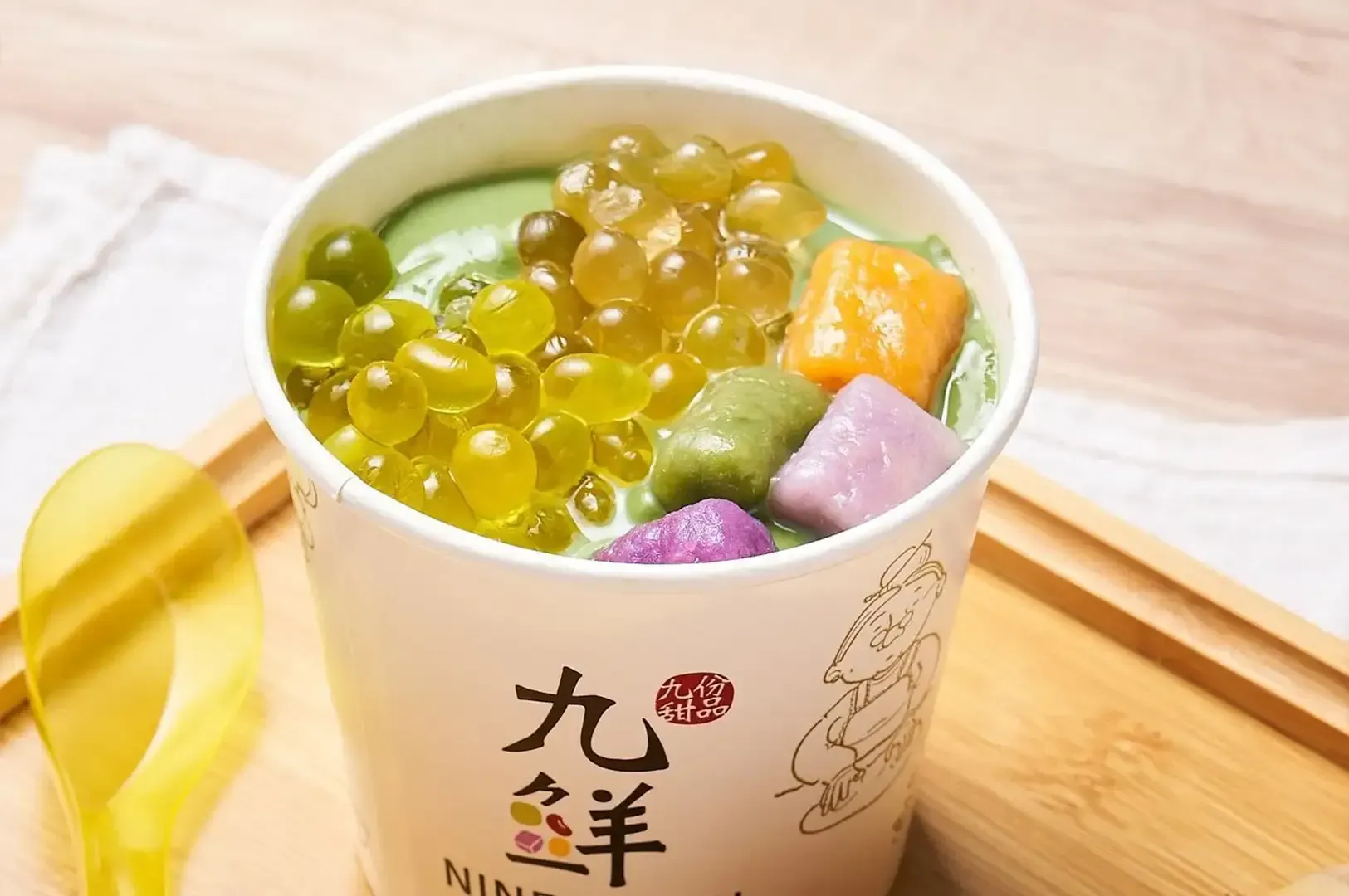 Cup of colorful shaved ice dessert with green syrup and fruit cubes on a wooden tray