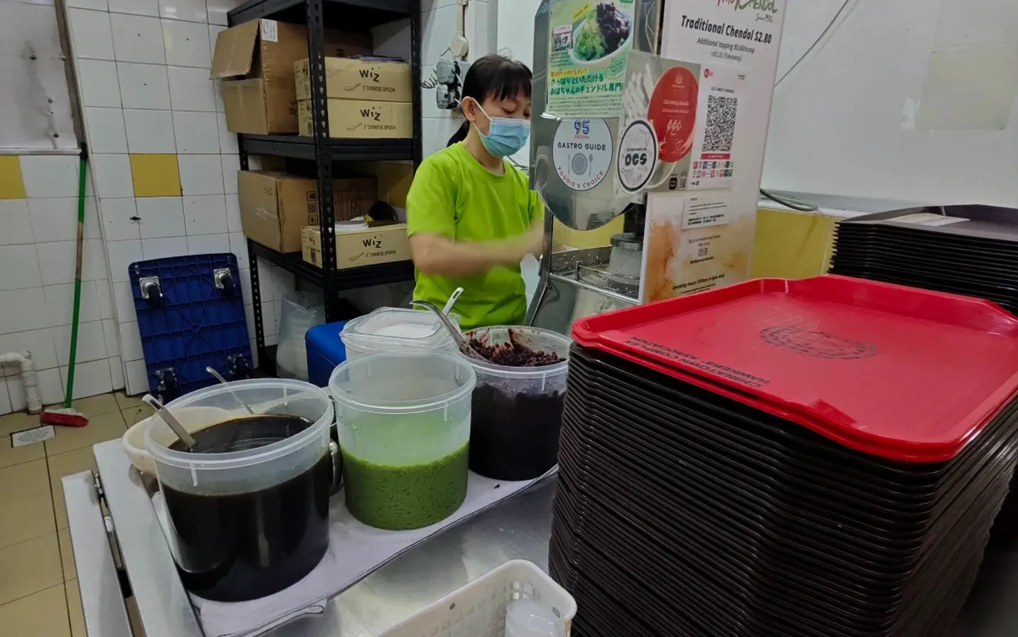 Person in a mask arranging large beverage containers and stacked trays at a counter.