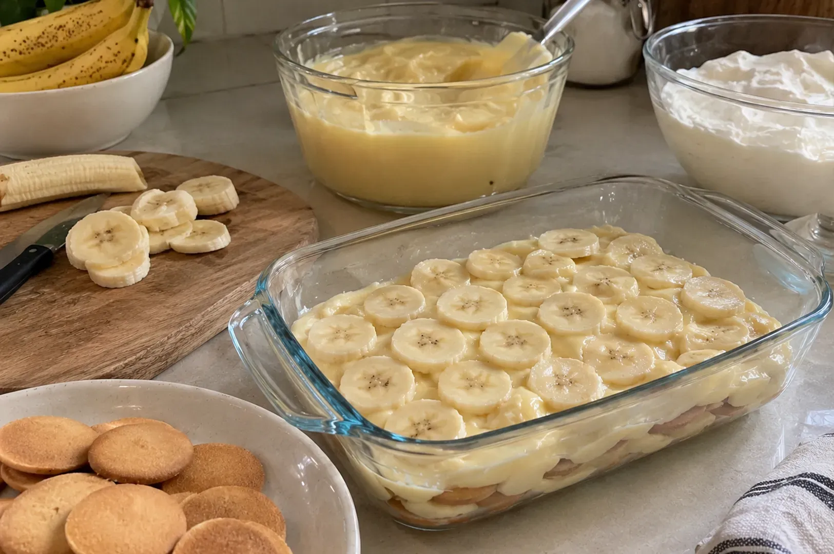 Banana pudding ingredients on a counter, with sliced bananas, wafers, and bowls of pudding and whipped cream
