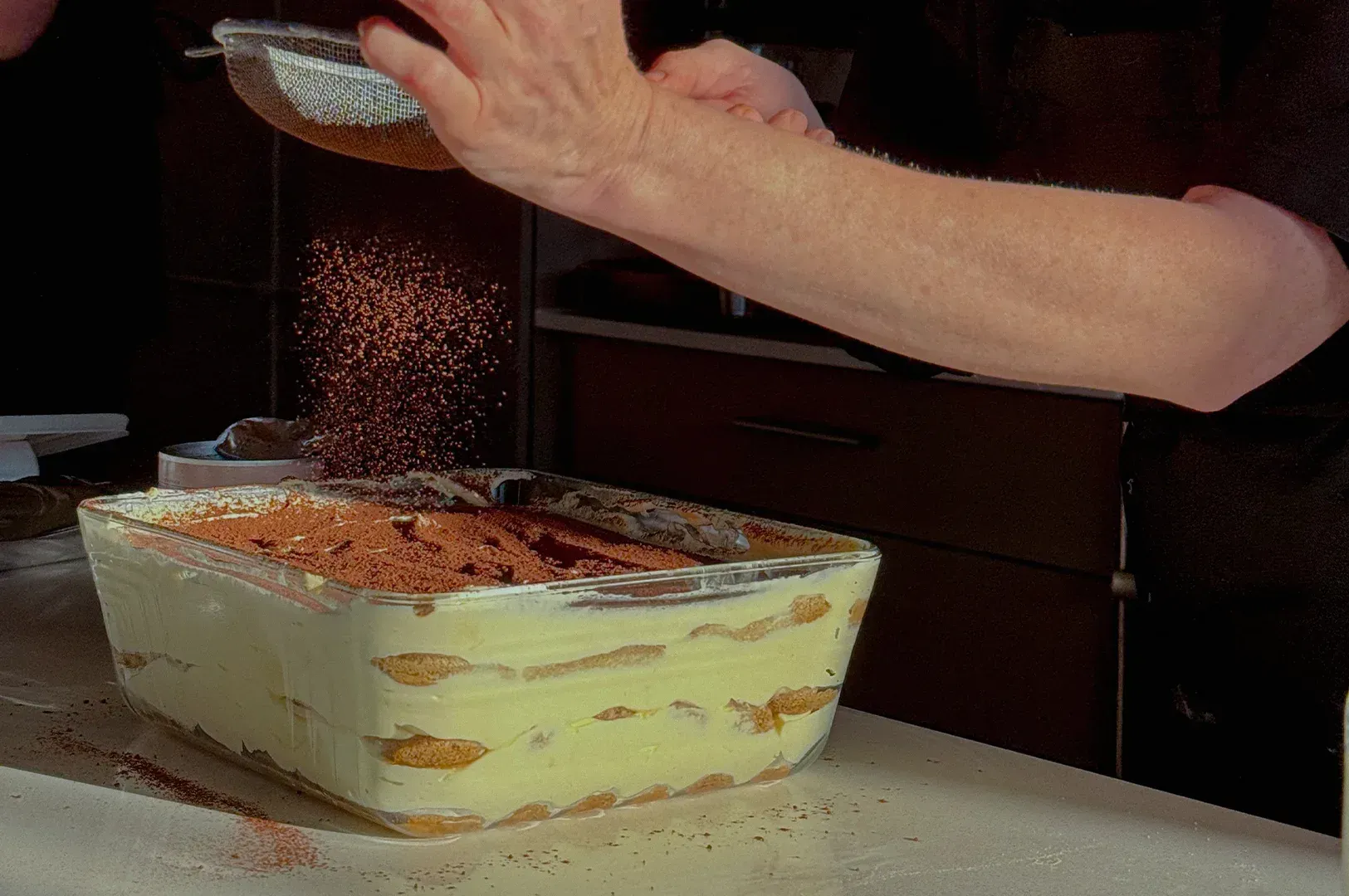 Hands sprinkle cocoa powder over a layered tiramisu in a glass dish