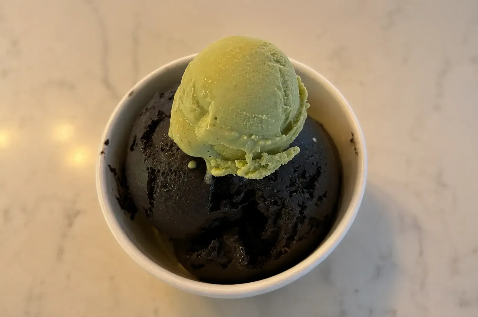 Cup of dark dessert topped with a pale green scoop, on a light marble table.