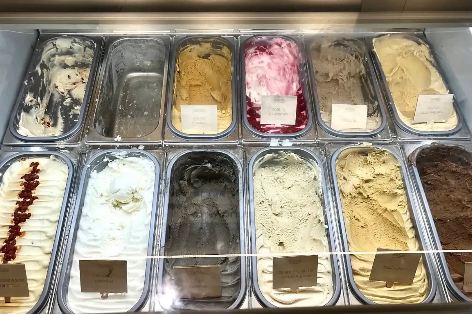 Ice cream display case with assorted flavors in metal tubs, including pink, beige, white, and dark chocolate.