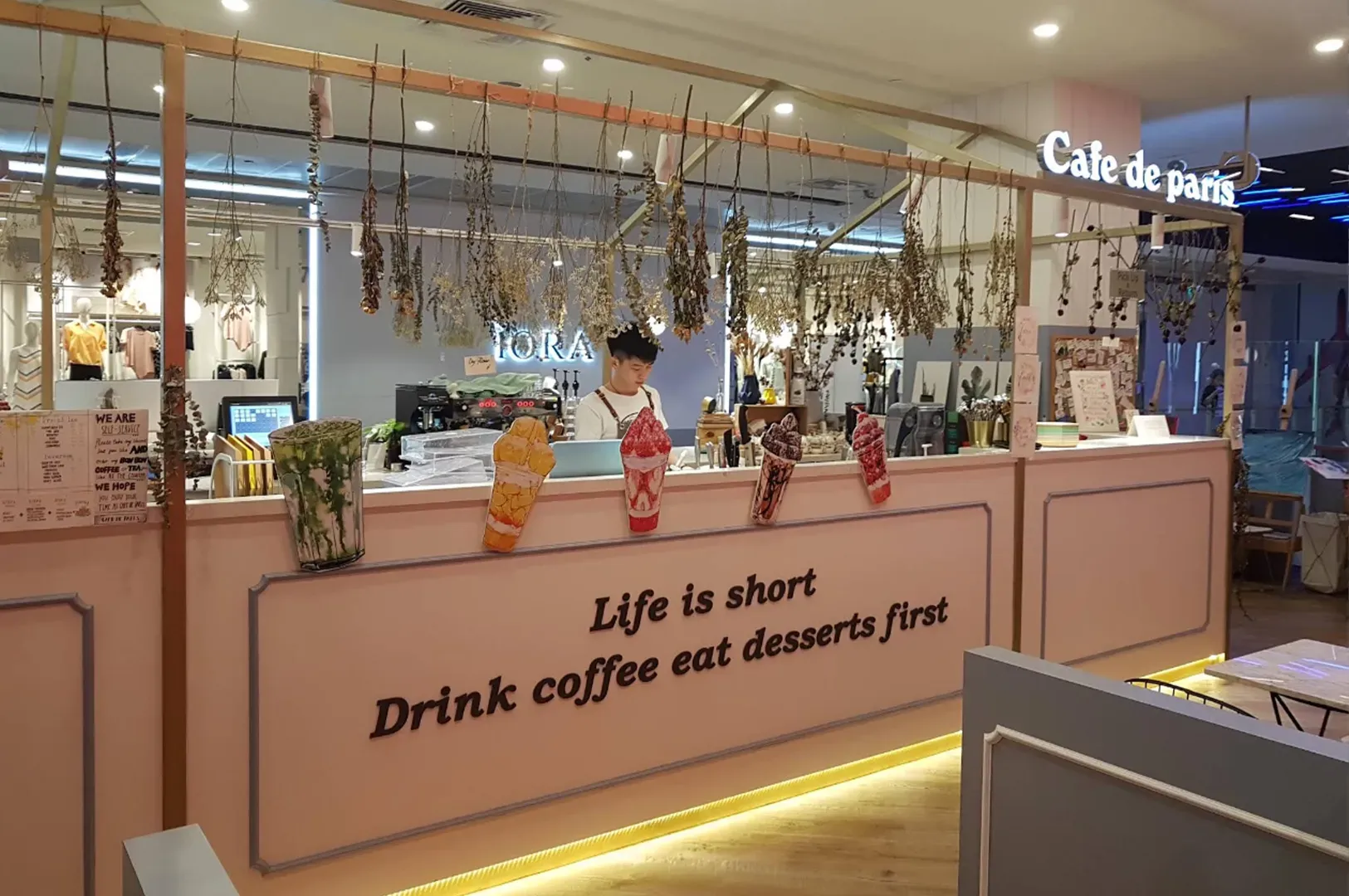 Cafe counter with a barista, string lights, and a sign reading “Life is short. Drink coffee and dessert first.”