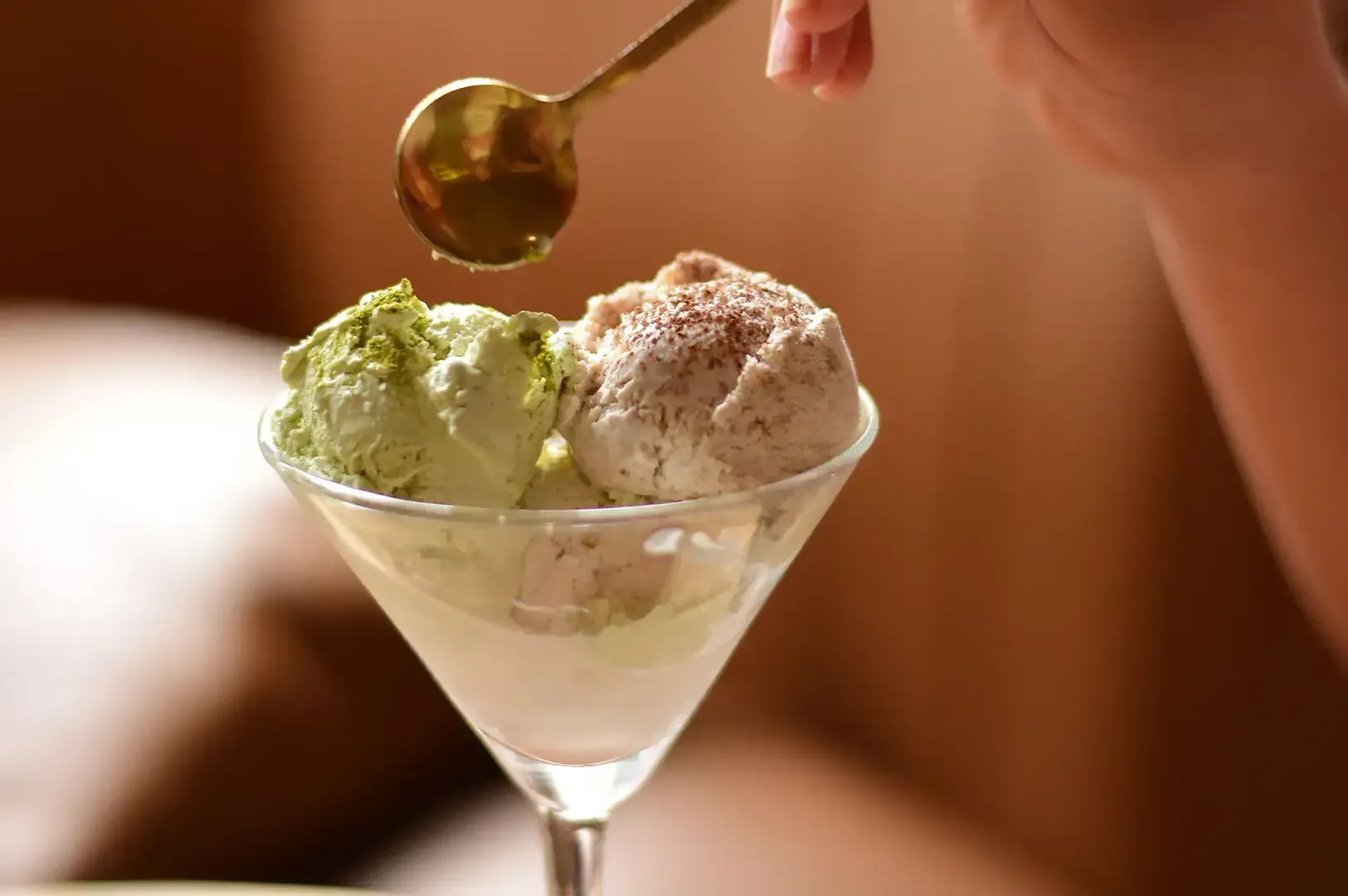 Hand placing a spoon over a martini glass with two scoops of green and beige ice cream