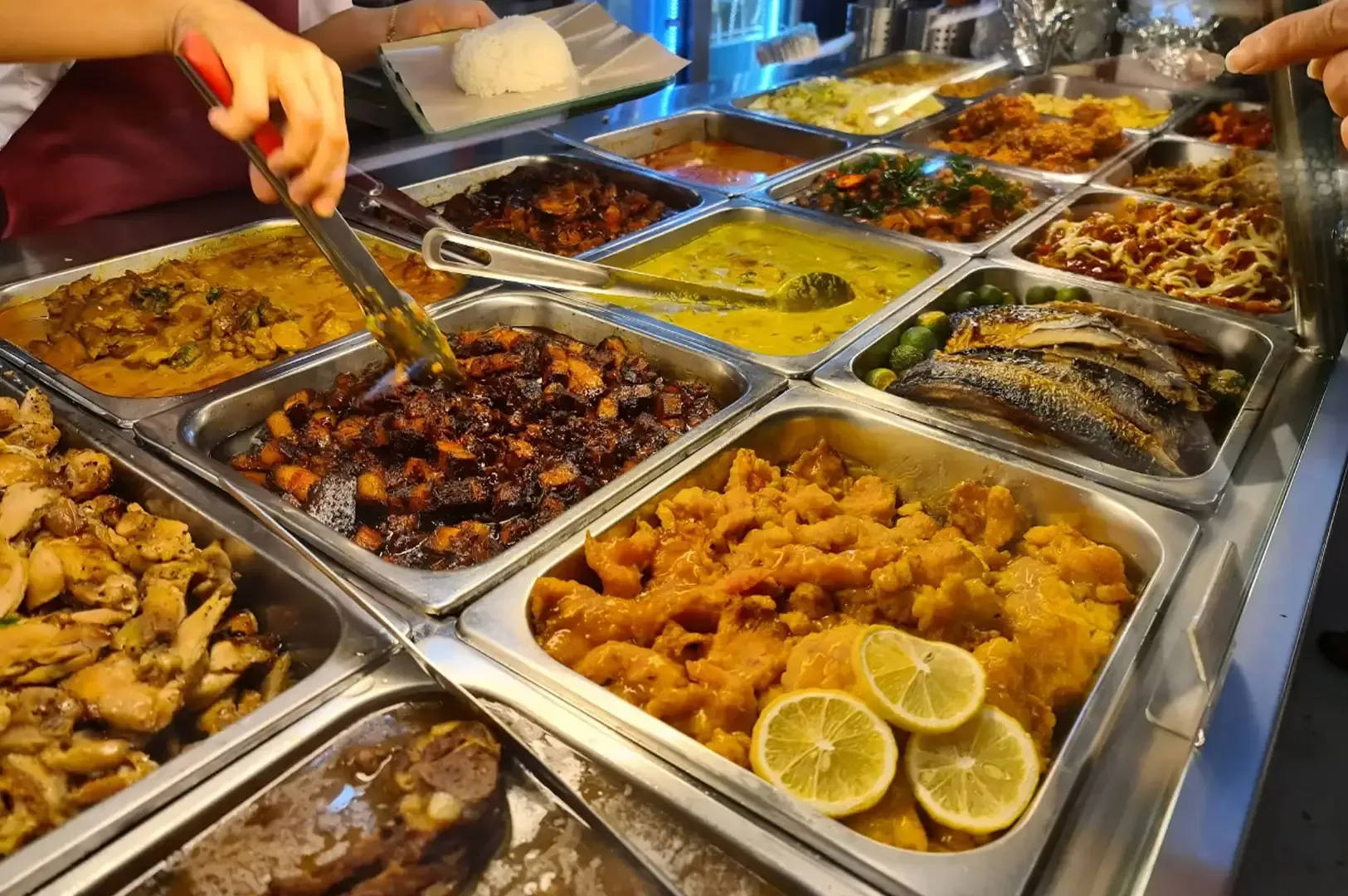 Buffet counter with trays of assorted cooked dishes, including grilled seafood and lemon slices, with a hand serving food