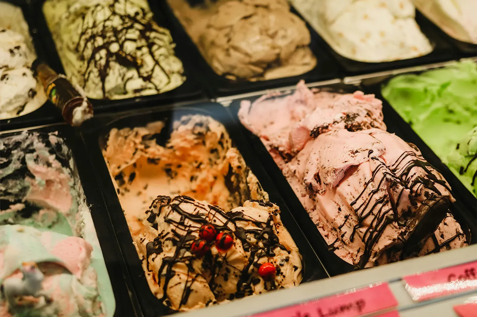 Assorted gelato flavors in a display case, including chocolate, green, and pink scoops with toppings