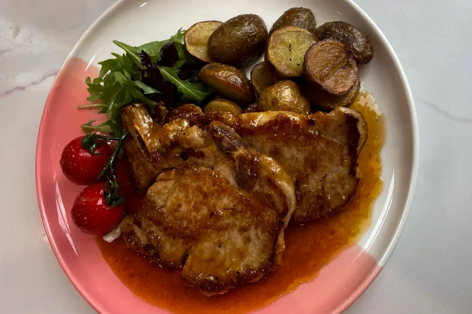 Plated roasted chicken with potatoes, cherry tomatoes, and greens on a pink-rimmed plate