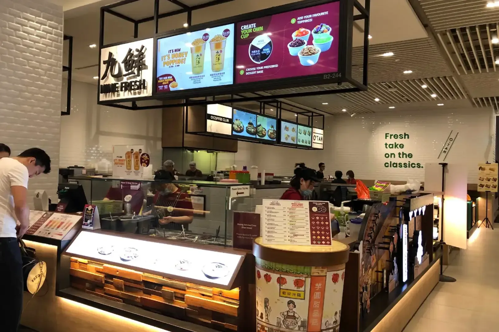 Fast-food counter with illuminated menu boards, staff behind the counter, and a customer ordering in a bright café.