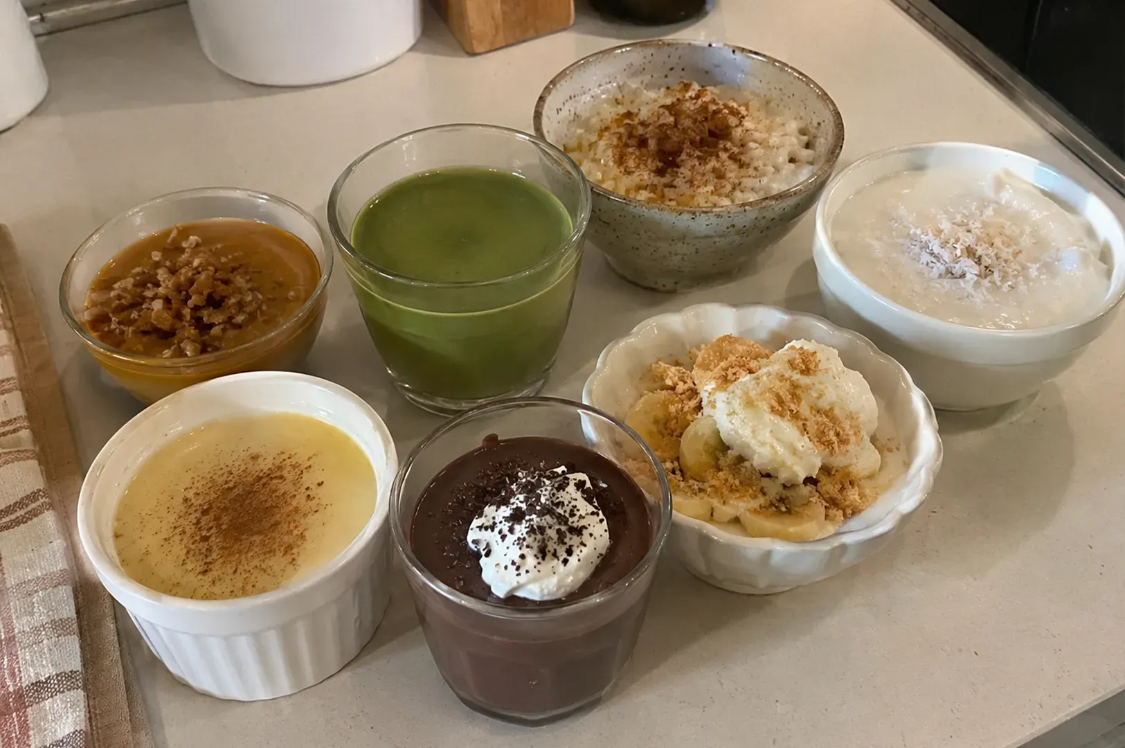 Assorted small dessert cups on a countertop, including green, chocolate, cream, and caramel-topped puddings