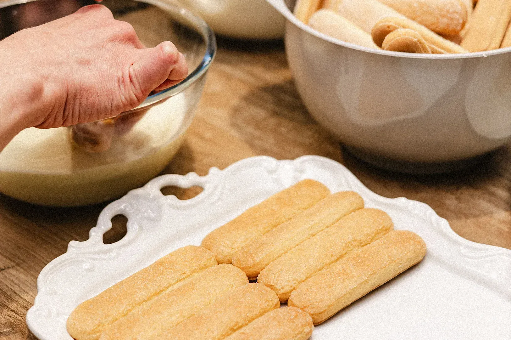 Hand shaping dough in a bowl beside trays of uncooked bread rolls on a table