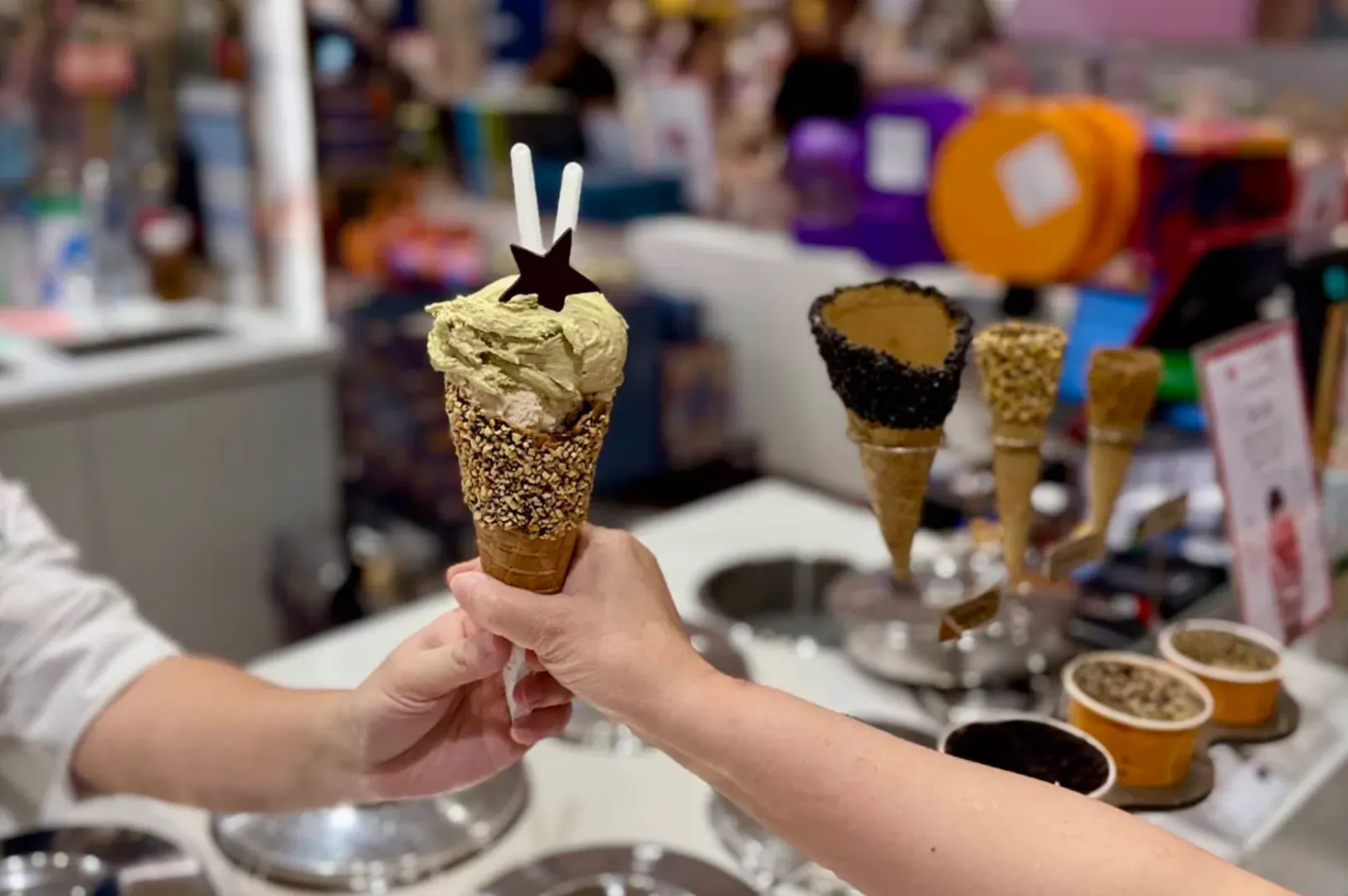 Two hands clink ice cream cones at a dessert stall with colorful toppings and cones in the background