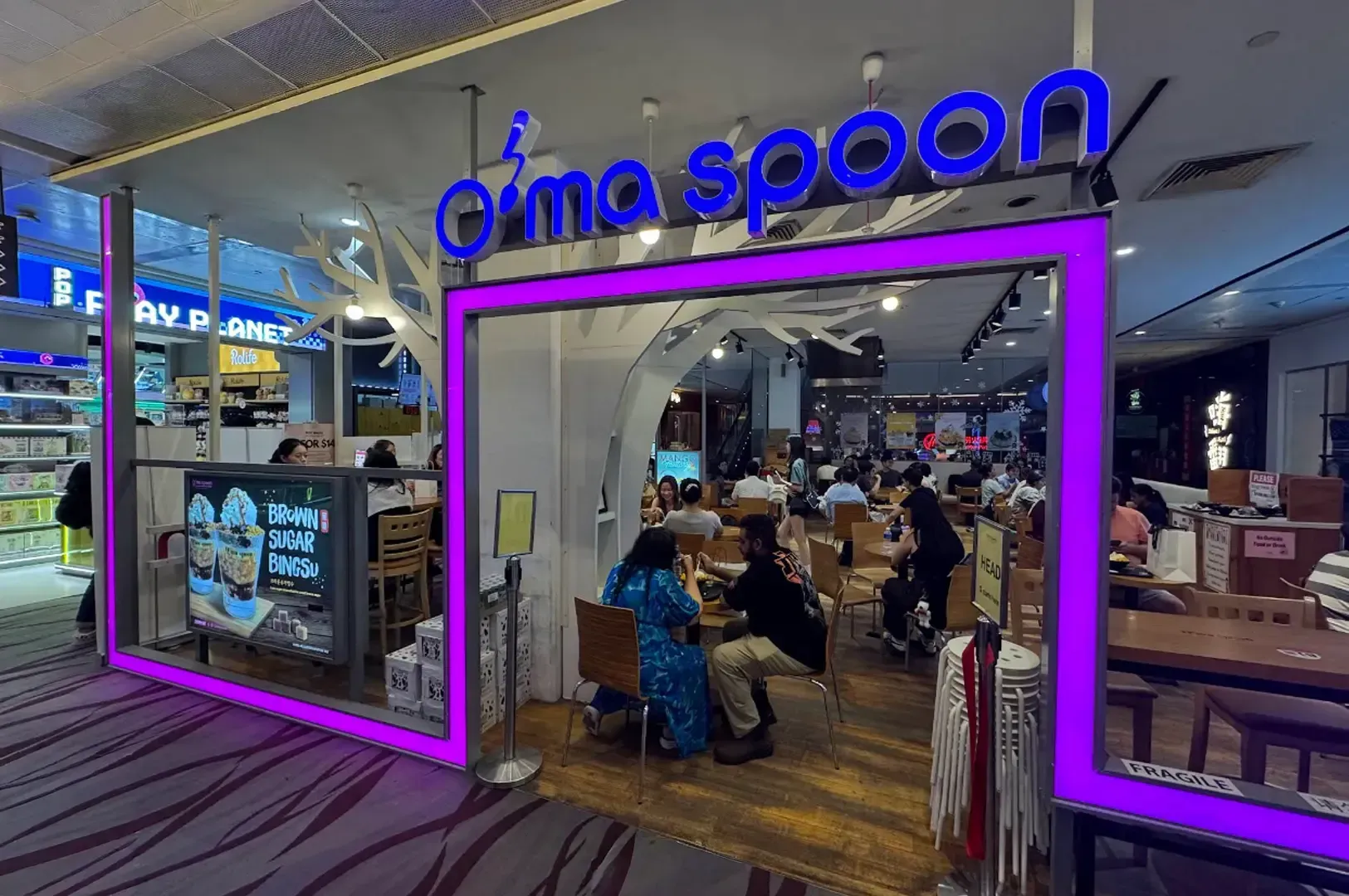 Oma Spoon restaurant entrance with purple neon frame and diners inside at a mall corridor