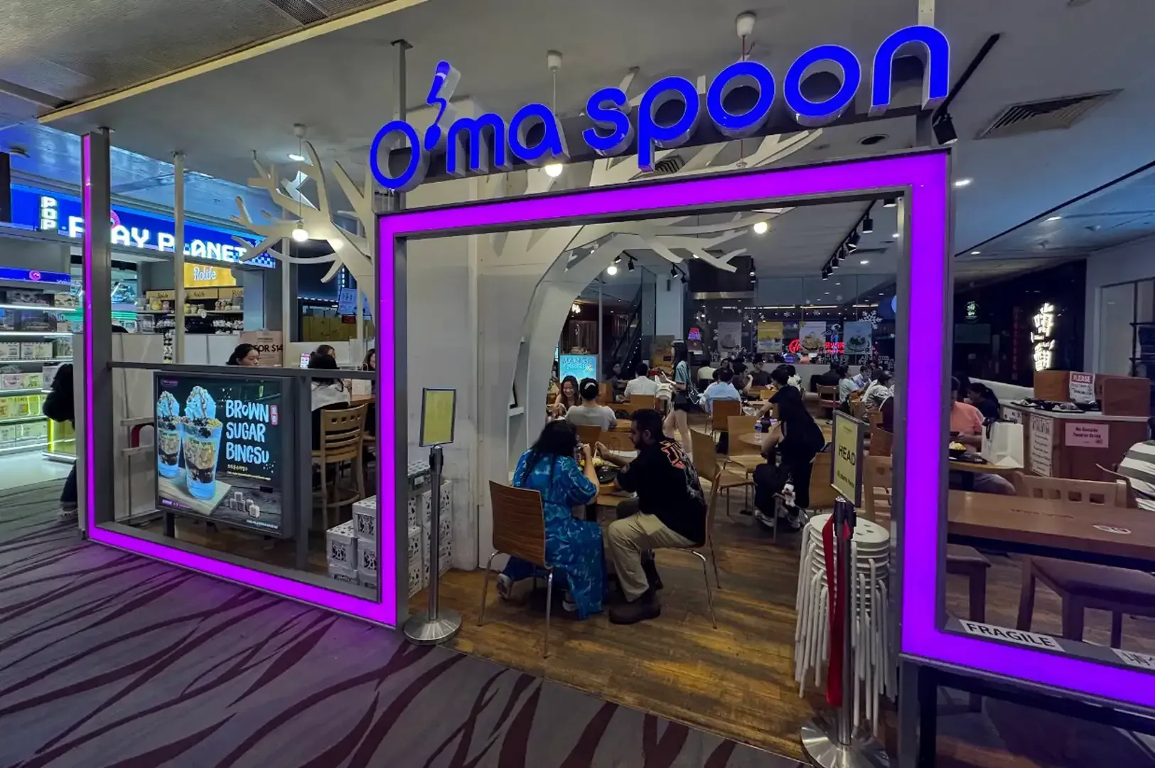 Entrance to Omo Spoon restaurant with neon sign and diners inside at a shopping mall