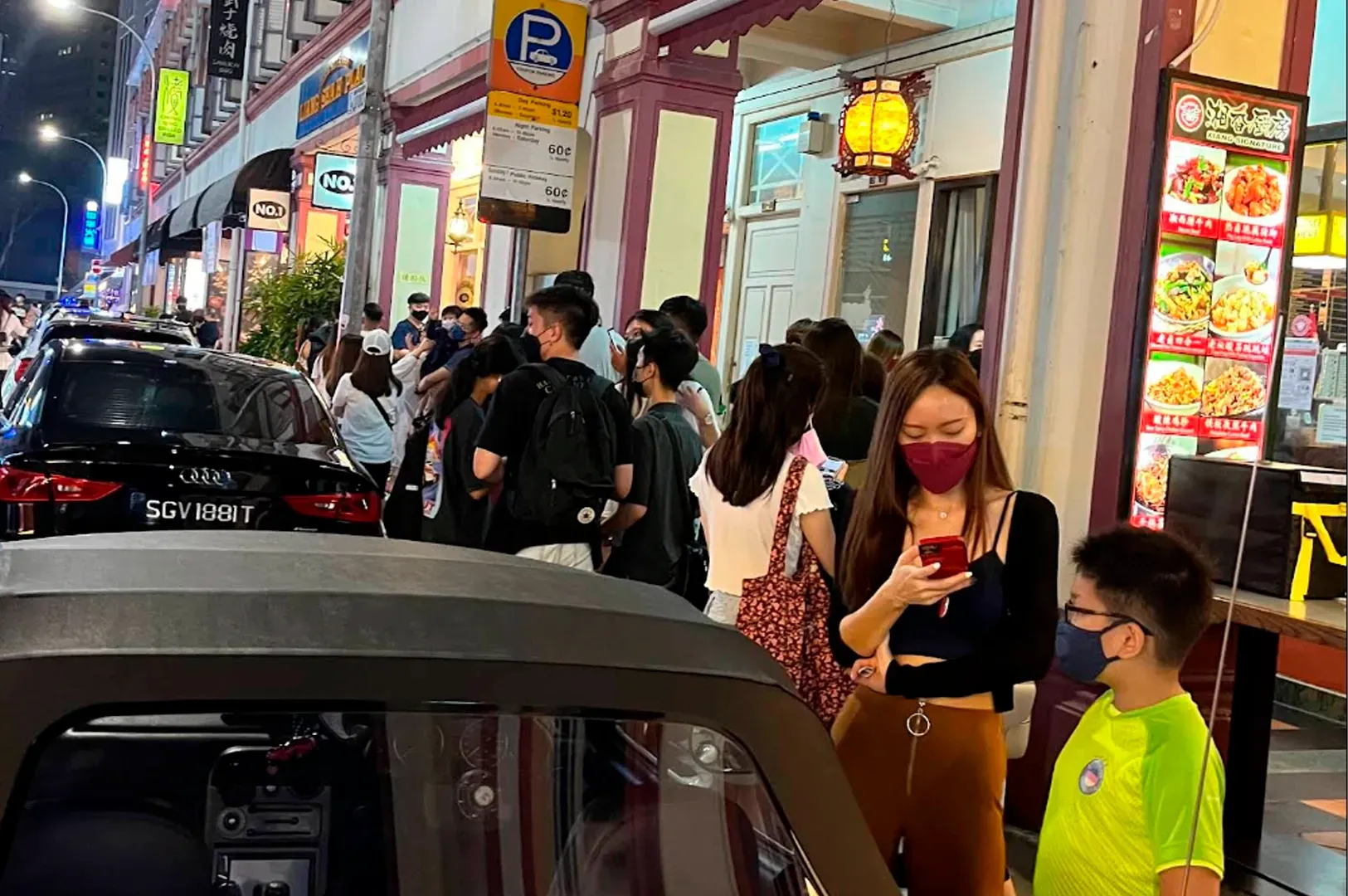 A night-time view of a long queue of people waiting along a sidewalk in front of colonial-style shophouses. Most people are looking at their phones or chatting while waiting to enter a restaurant. Several cars are parked along the curb, and a bright red digital menu board for a nearby eatery stands on the right.