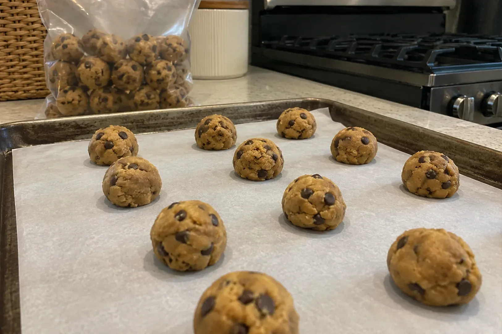 Cookie dough balls on parchment paper in a kitchen, with a bag of dough and a stove in the background