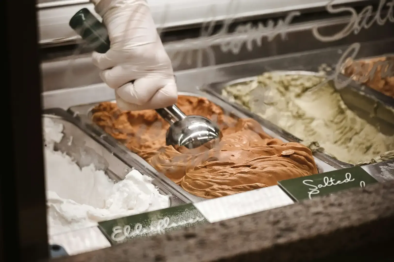 Hand scooping chocolate gelato from a display case with vanilla and other flavors nearby