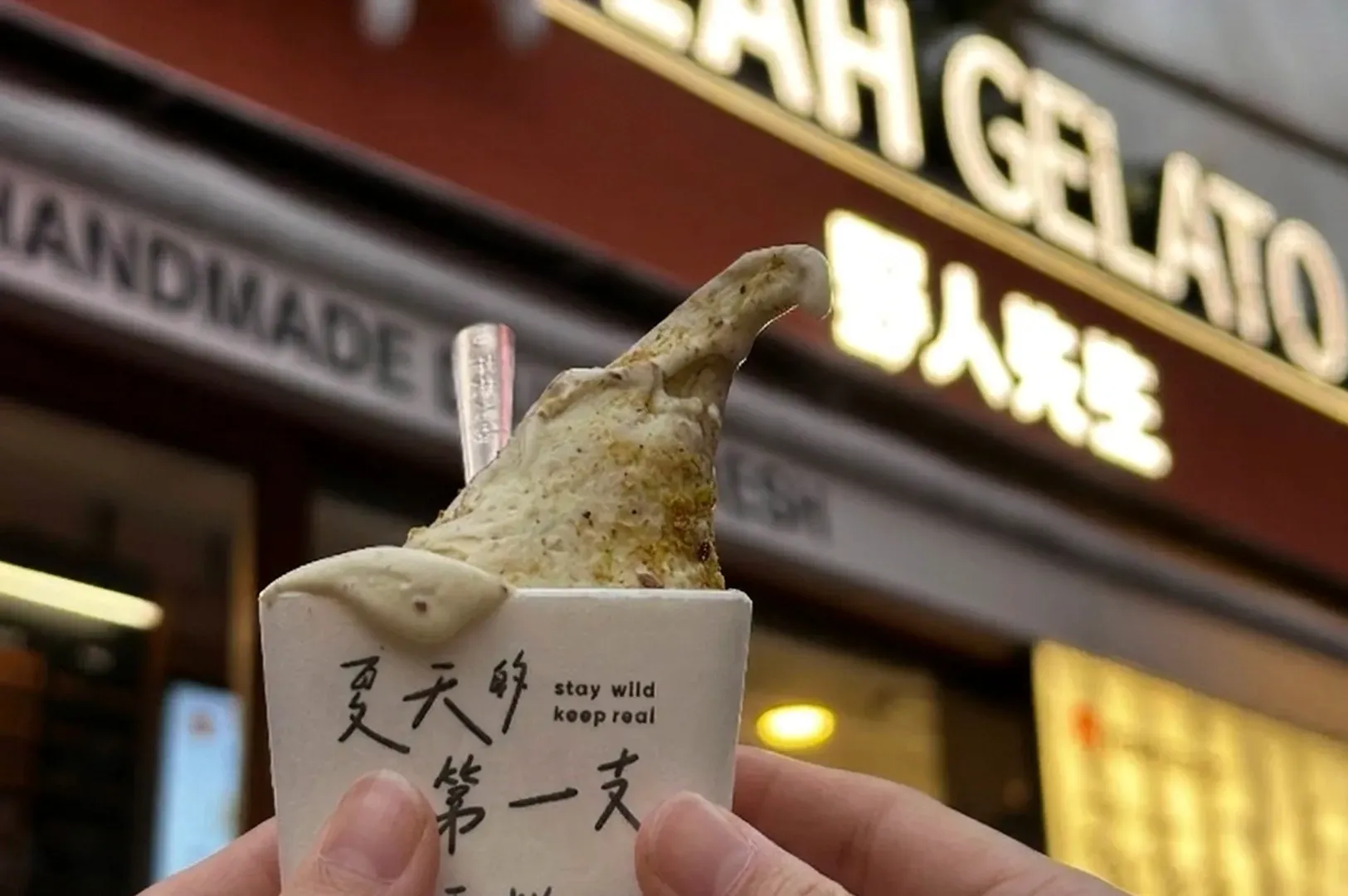 A close-up shot of a person holding a small paper cup containing a scoop of pistachio-colored gelato. The gelato is textured with small nut pieces. The background shows a blurry, warm-toned neon sign that reads