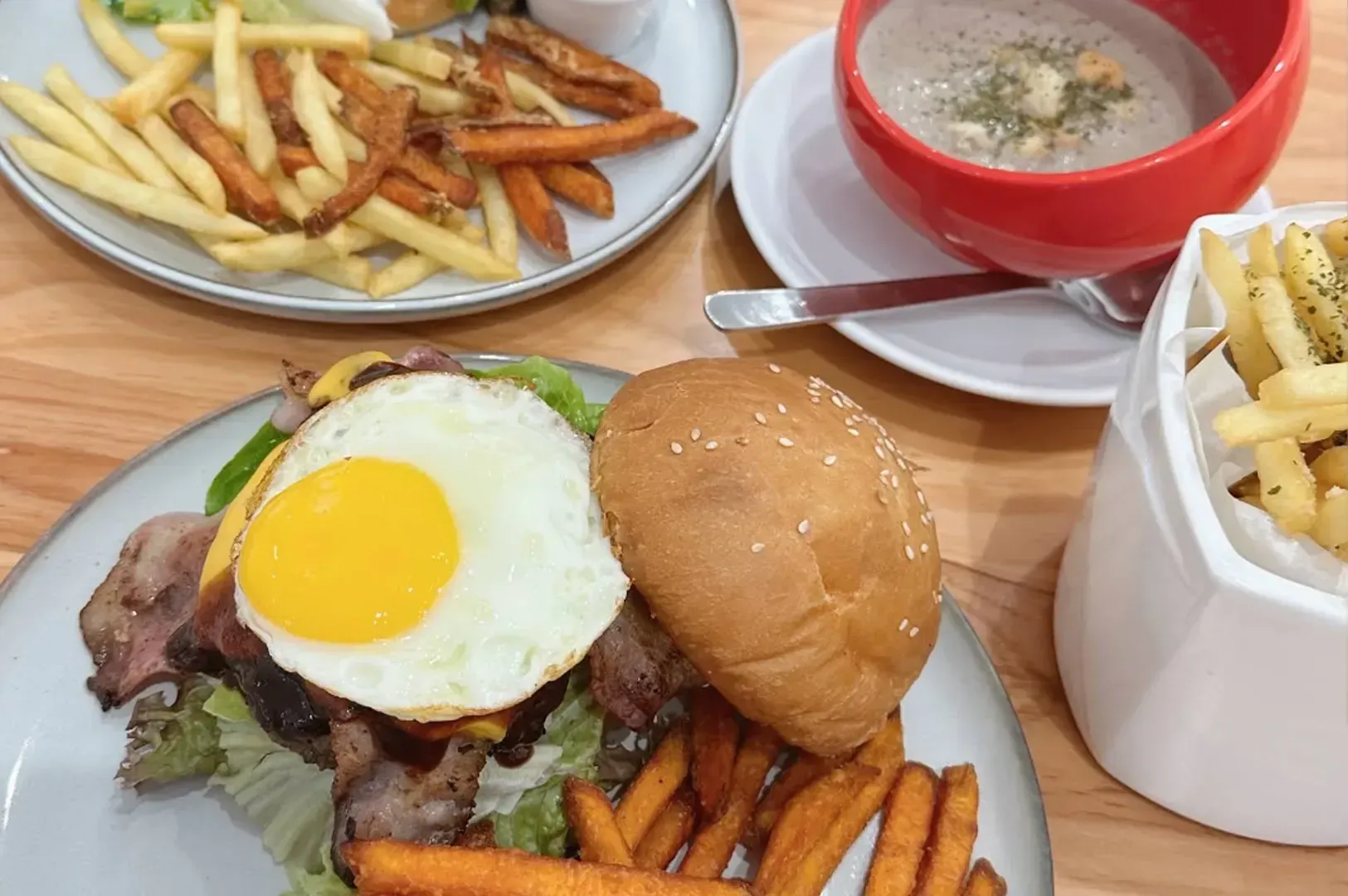 Burger, fries, and a fried egg on a plate beside soup and a carton of fries on a wooden table