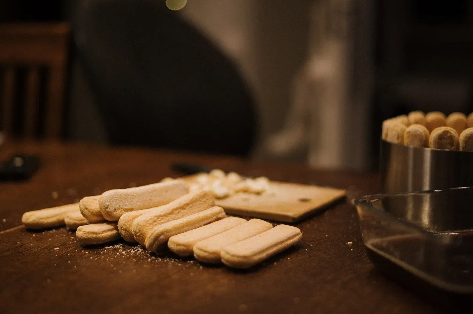 Ladyfingers on a dark table beside a metal cup, with crumbs and a blurred chair in the background