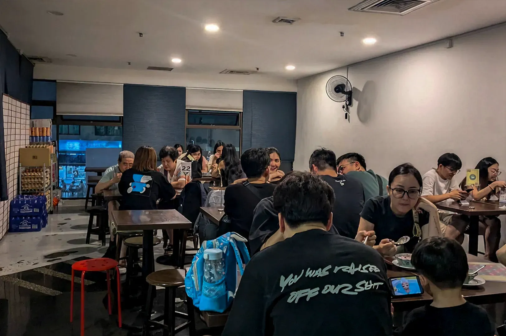 The bustling atmosphere inside the dessert parlor during peak hours. The room is filled with a diverse crowd of diners—families, friends, and couples—all huddled around small tables. The lighting is warm and functional, reflecting off the polished dark floors. In the corner, crates of bottled water and supplies are stacked high, reinforcing the casual,