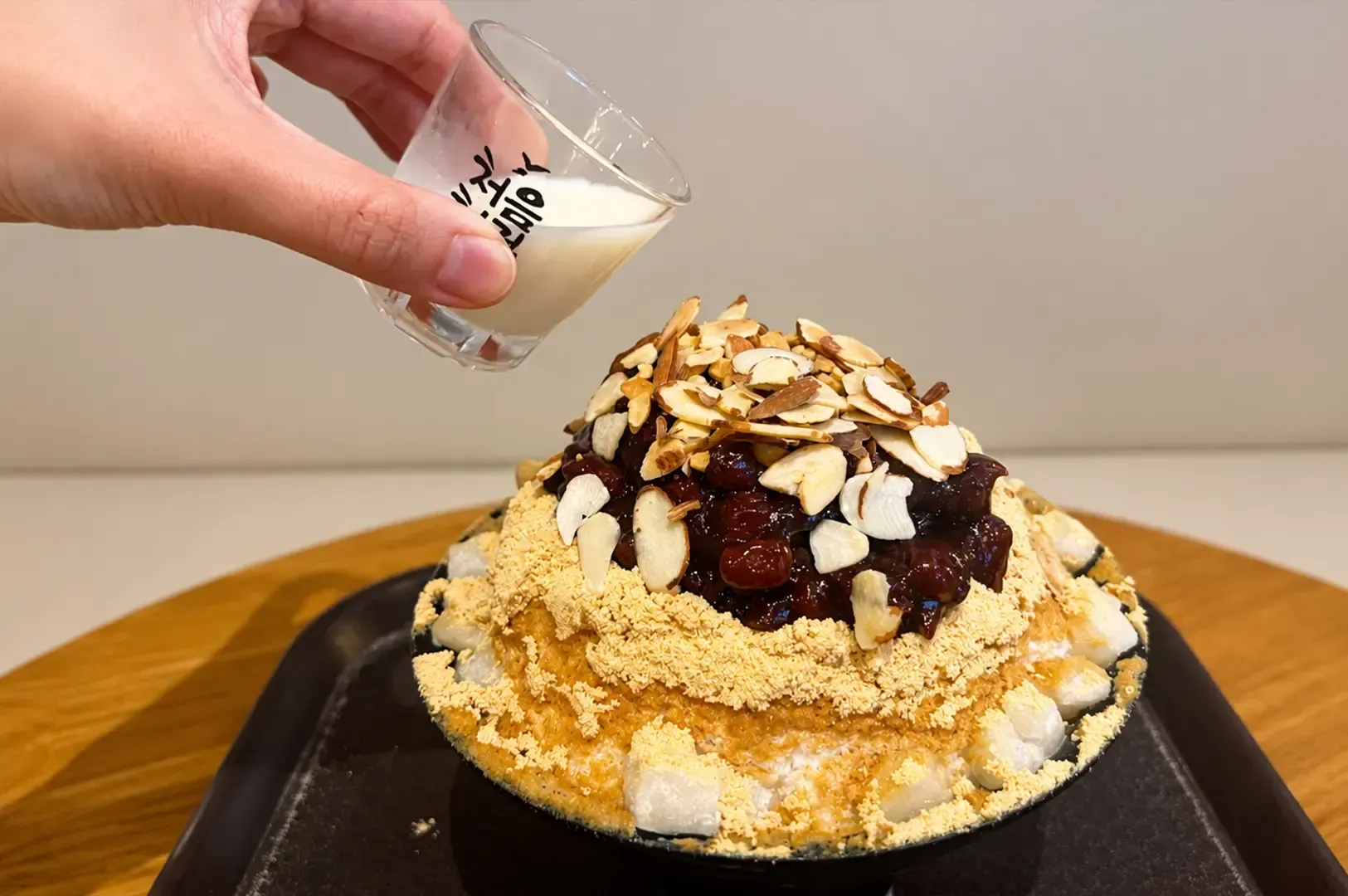 Hand pouring cream onto a shaved ice dessert topped with nuts and syrup on a tray
