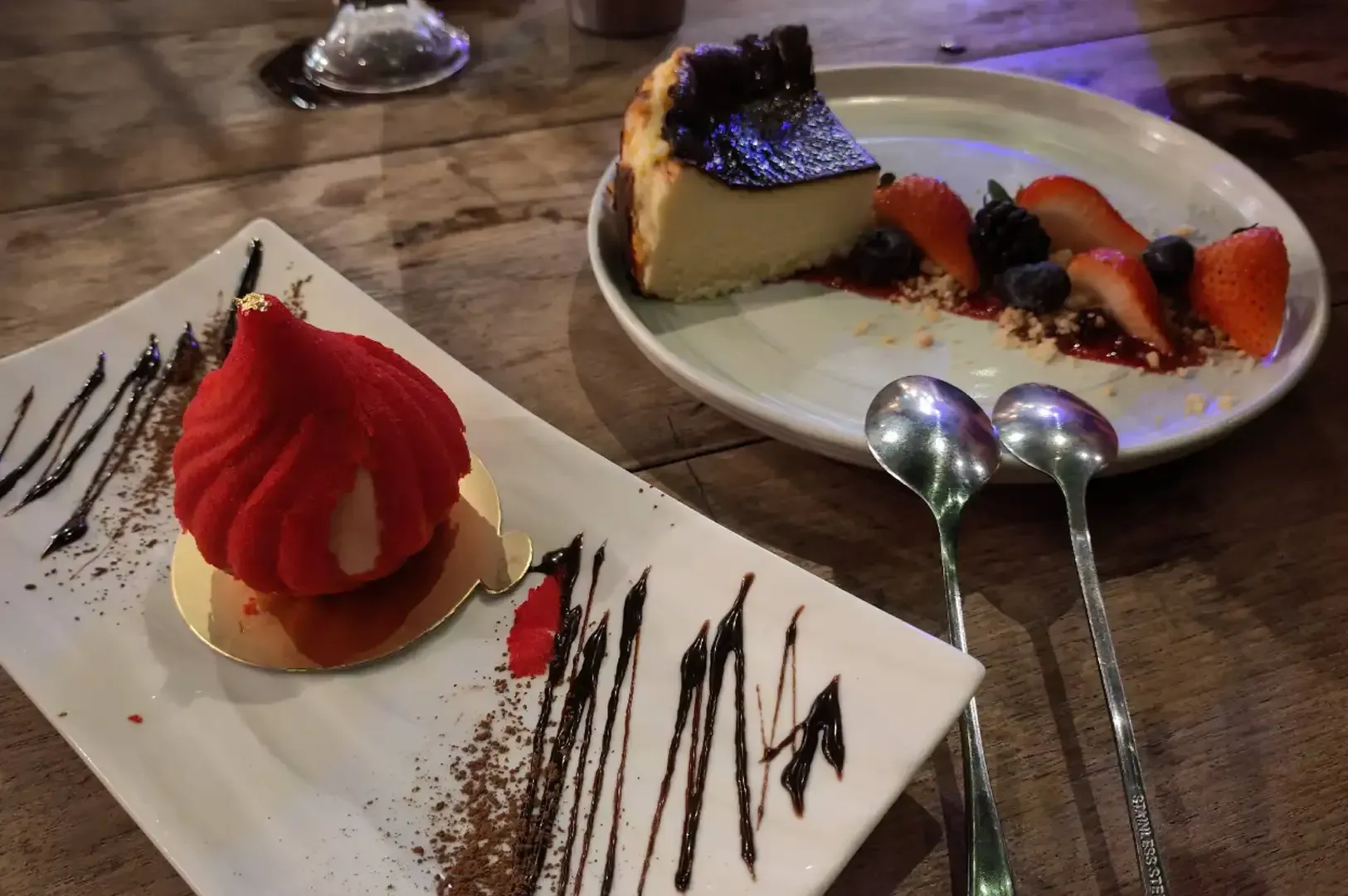 Desserts on a wooden table: red tart, cheesecake with berries, and two spoons on a plate.
