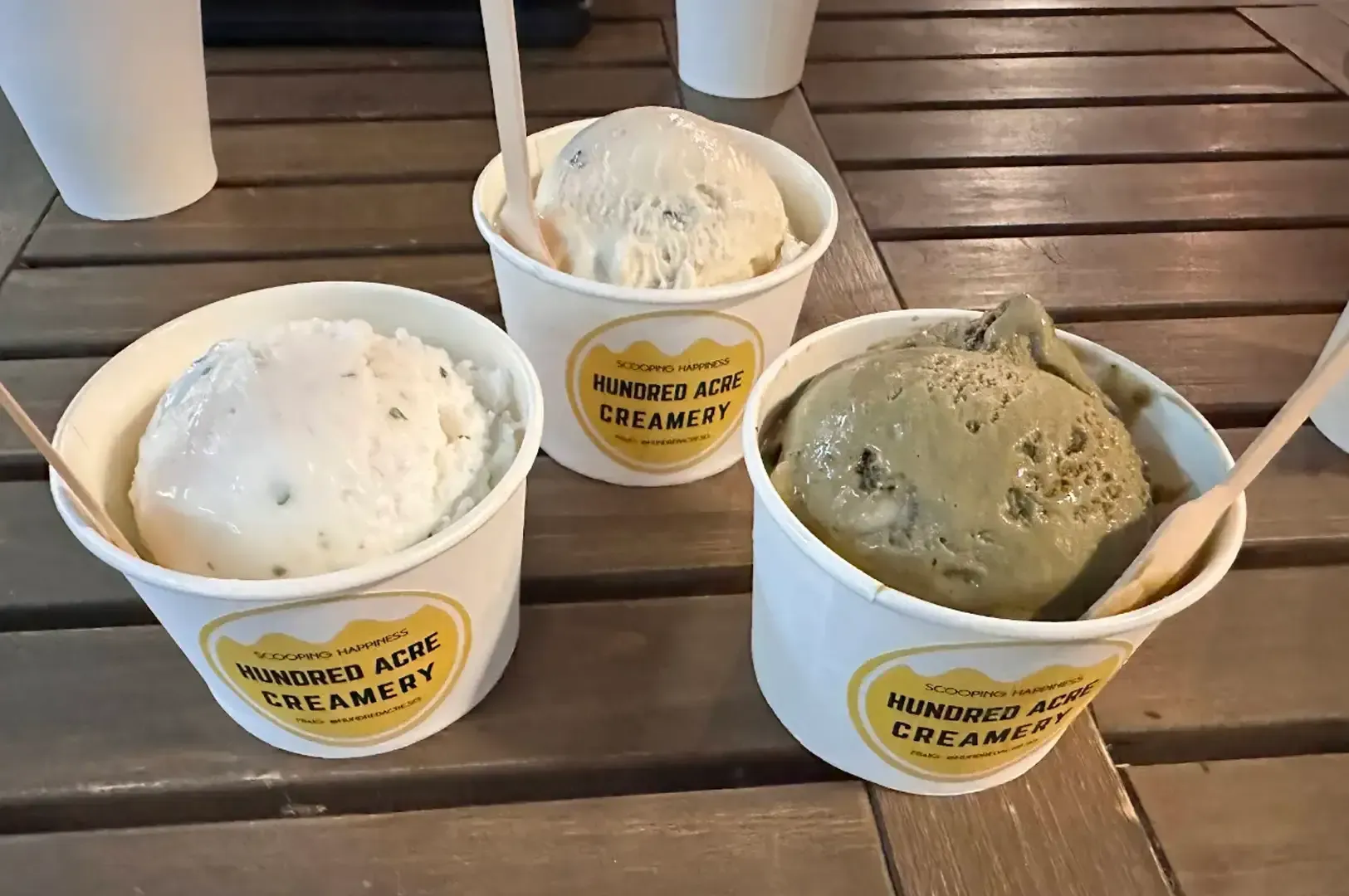 Three cups of ice cream on a wooden table, including white and green scoops with spoons.