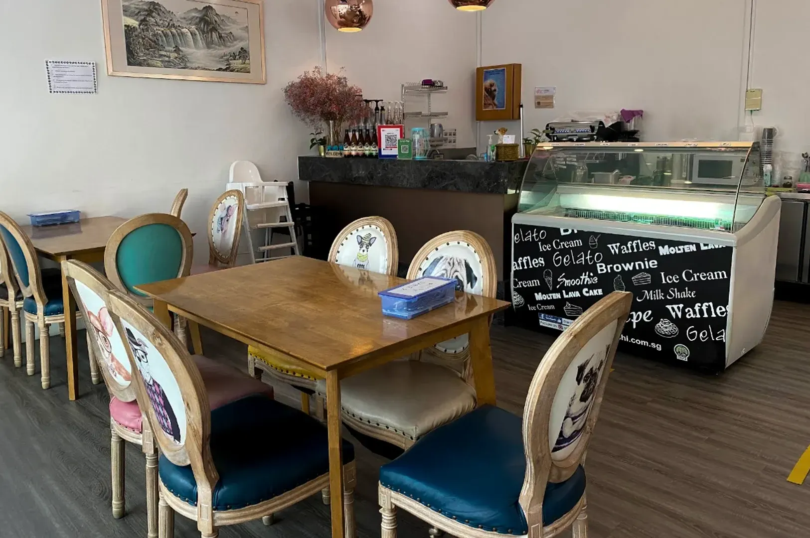 Cozy café with wooden tables, teal chairs, and a glass display counter with a chalkboard menu.