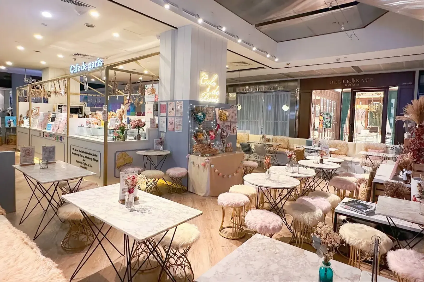 Bright café interior with marble tables, wicker chairs, and a colorful dessert counter beneath warm lighting