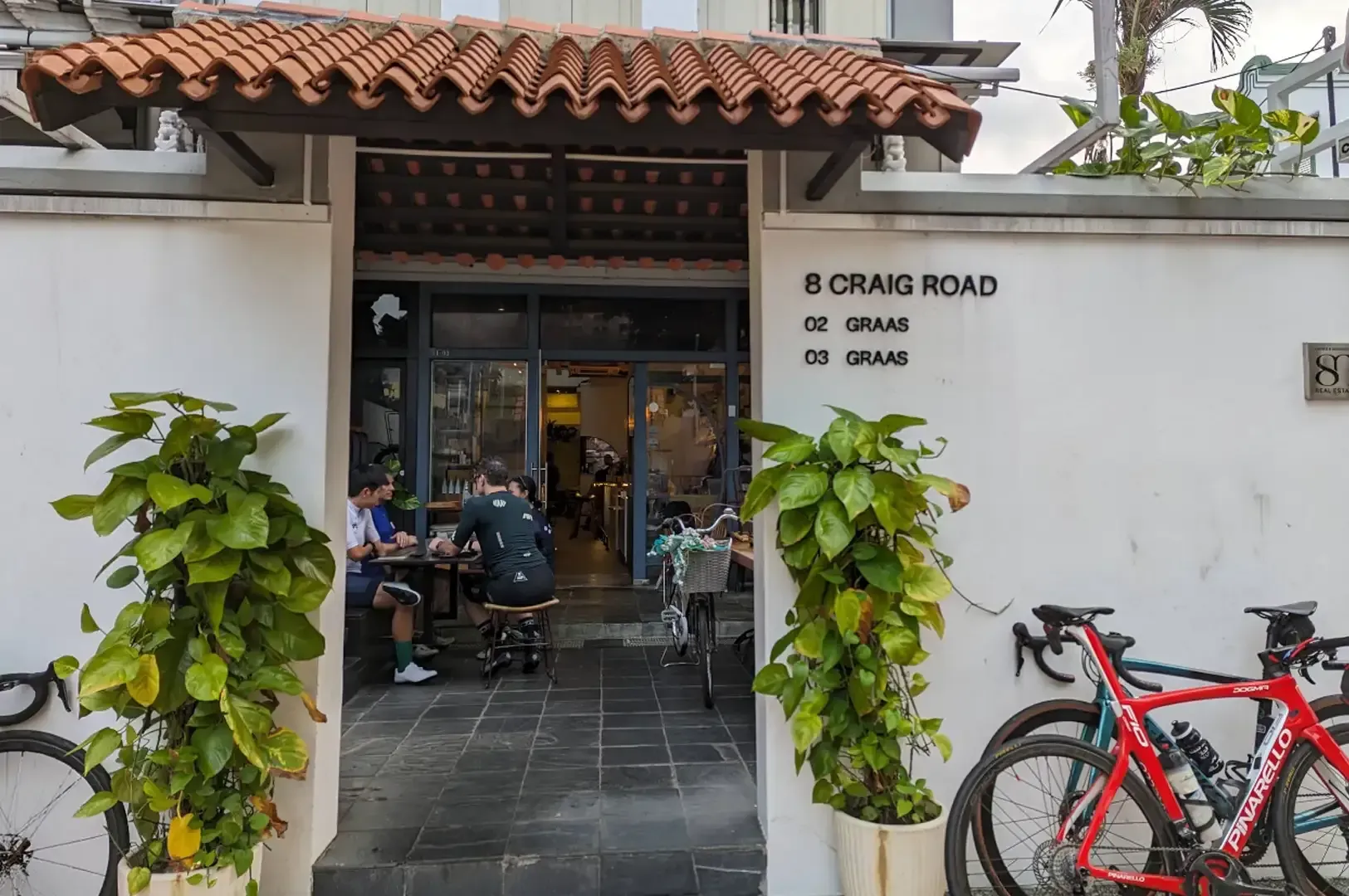 Cafe entrance with terracotta roof, potted plants, and a red bicycle beside a white wall sign reading “S. CRAIG ROAD.”