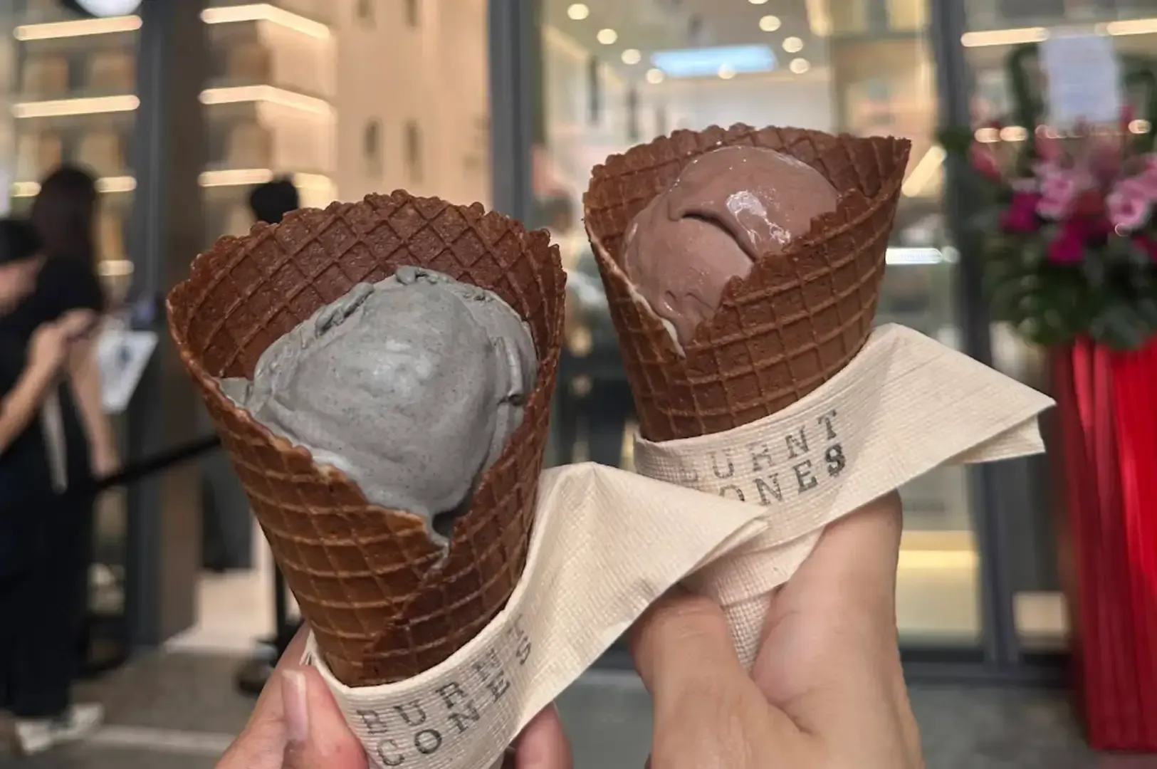 Two waffle cones with gray and brown ice cream held up in a shop