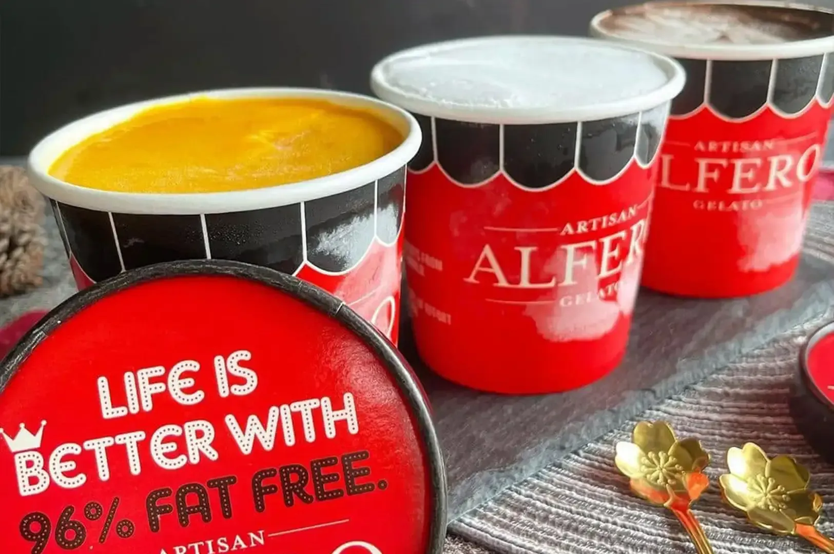 Red ALFERO cups on a table, one open with yellow drink; lid reads “Life is better with 96% fat free.”
