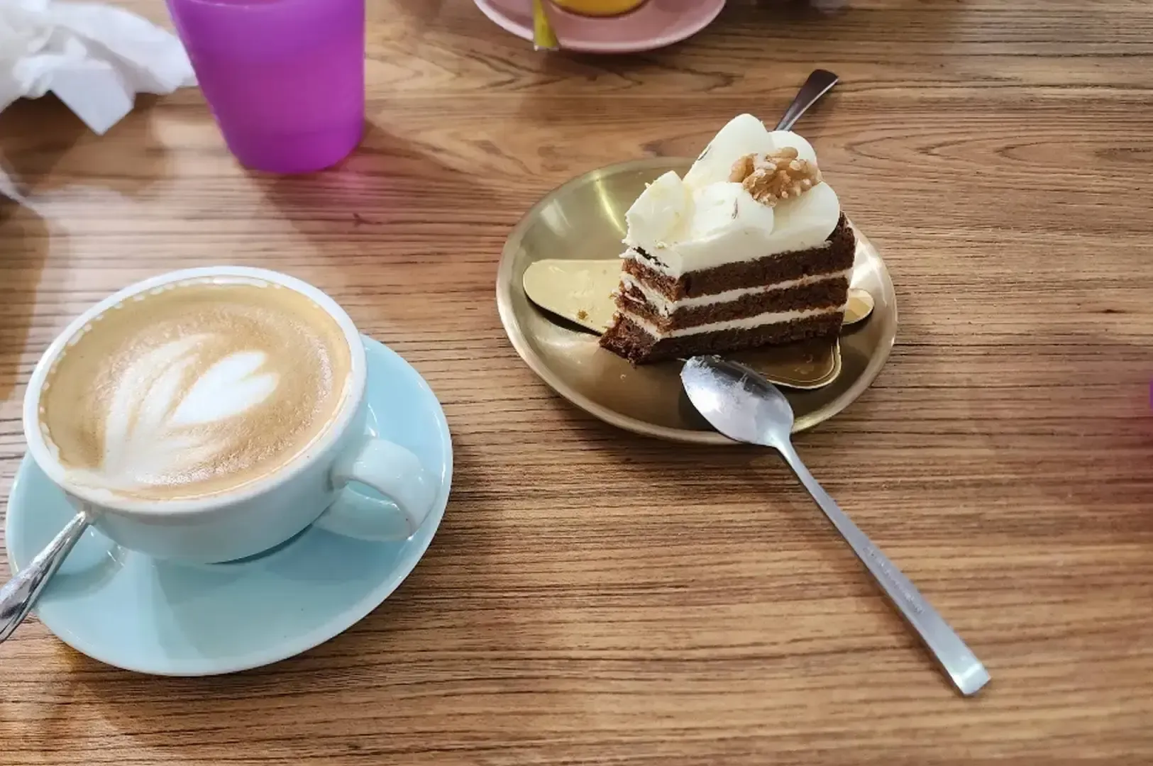 Coffee and a slice of chocolate cake on a wooden table, with a spoon and purple cup nearby.