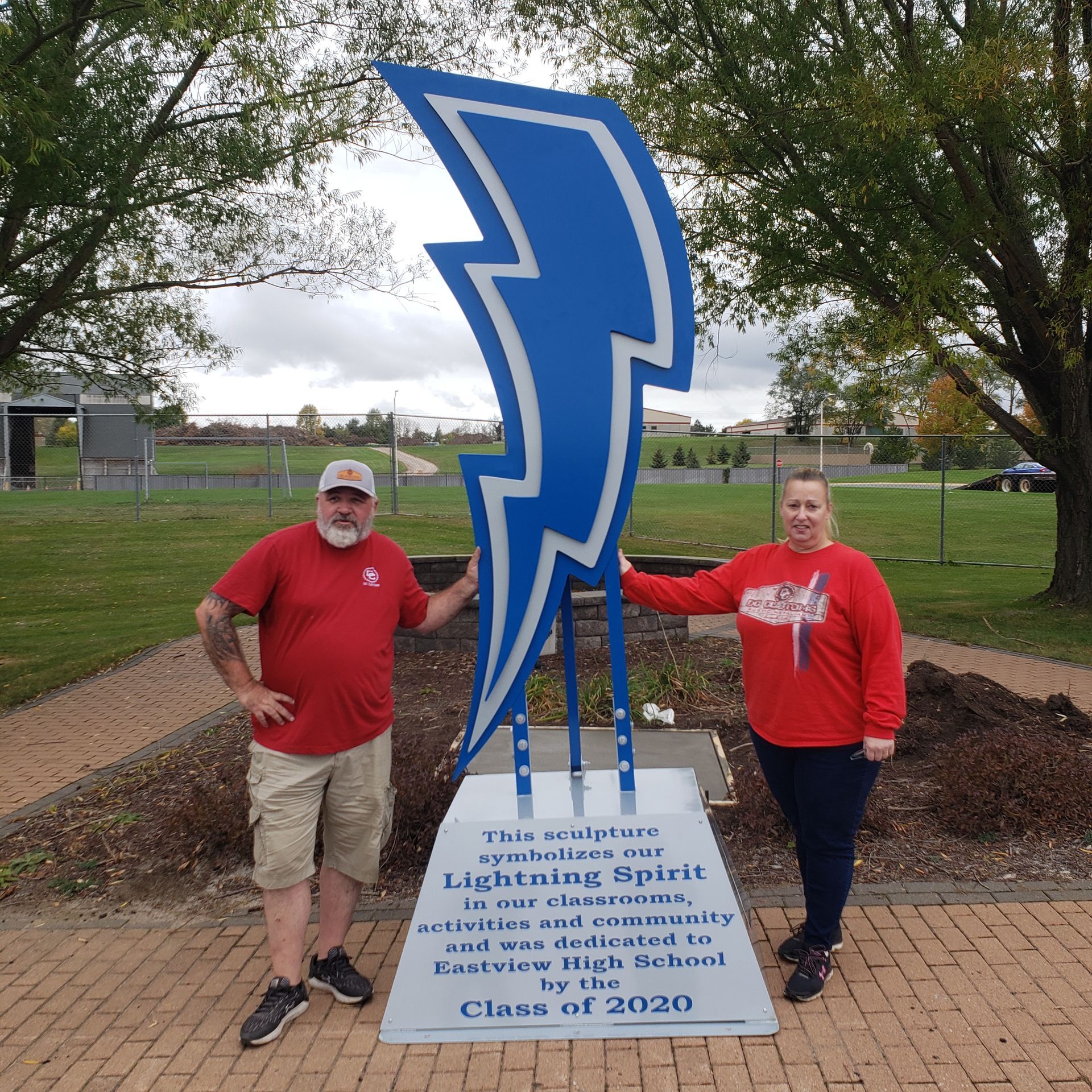 Two people stand by a blue lightning bolt sculpture in front of a school.