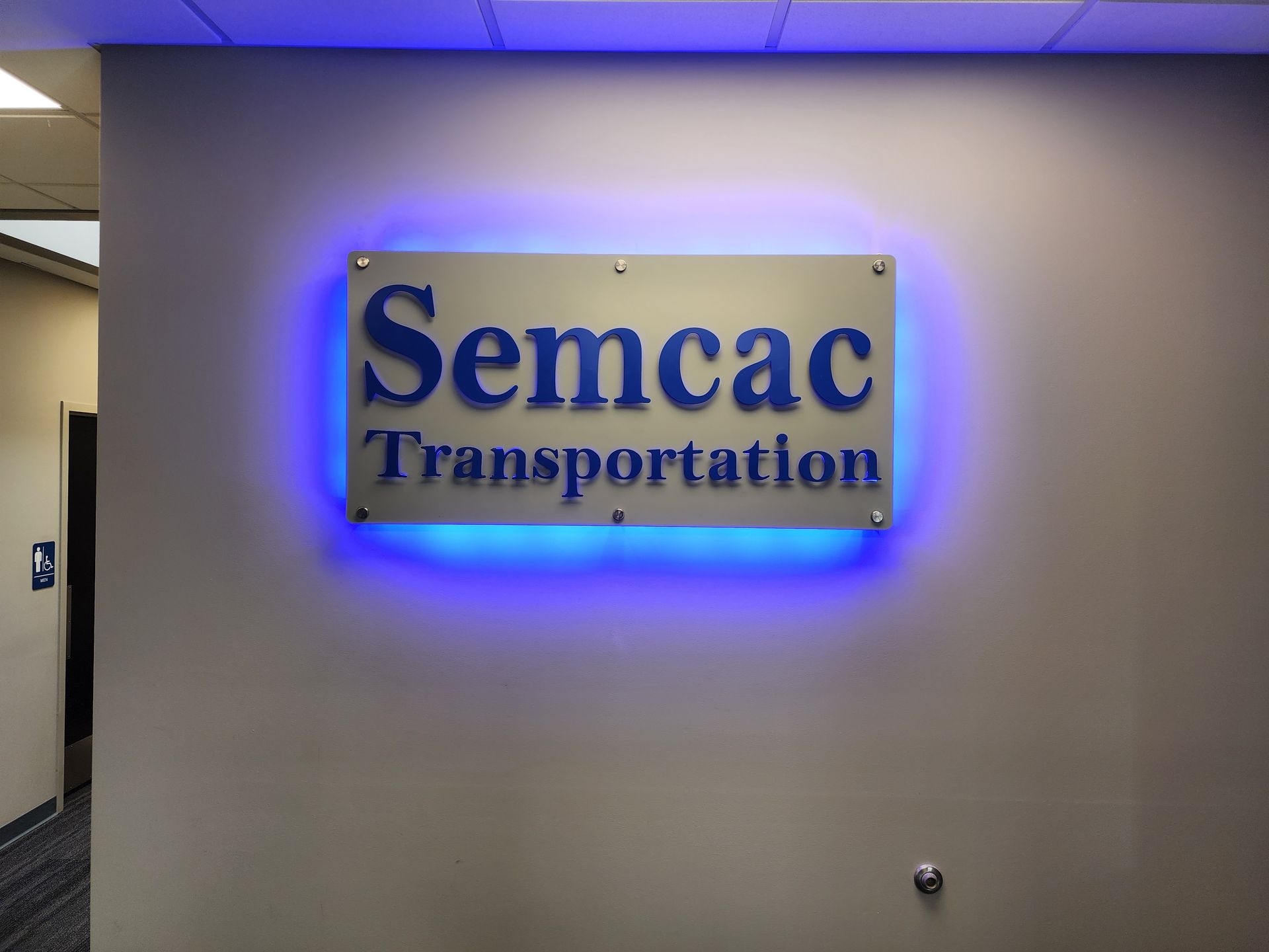 Semcac Transportation sign with blue text on a metallic background, lit by a blue halo, mounted on a beige wall.