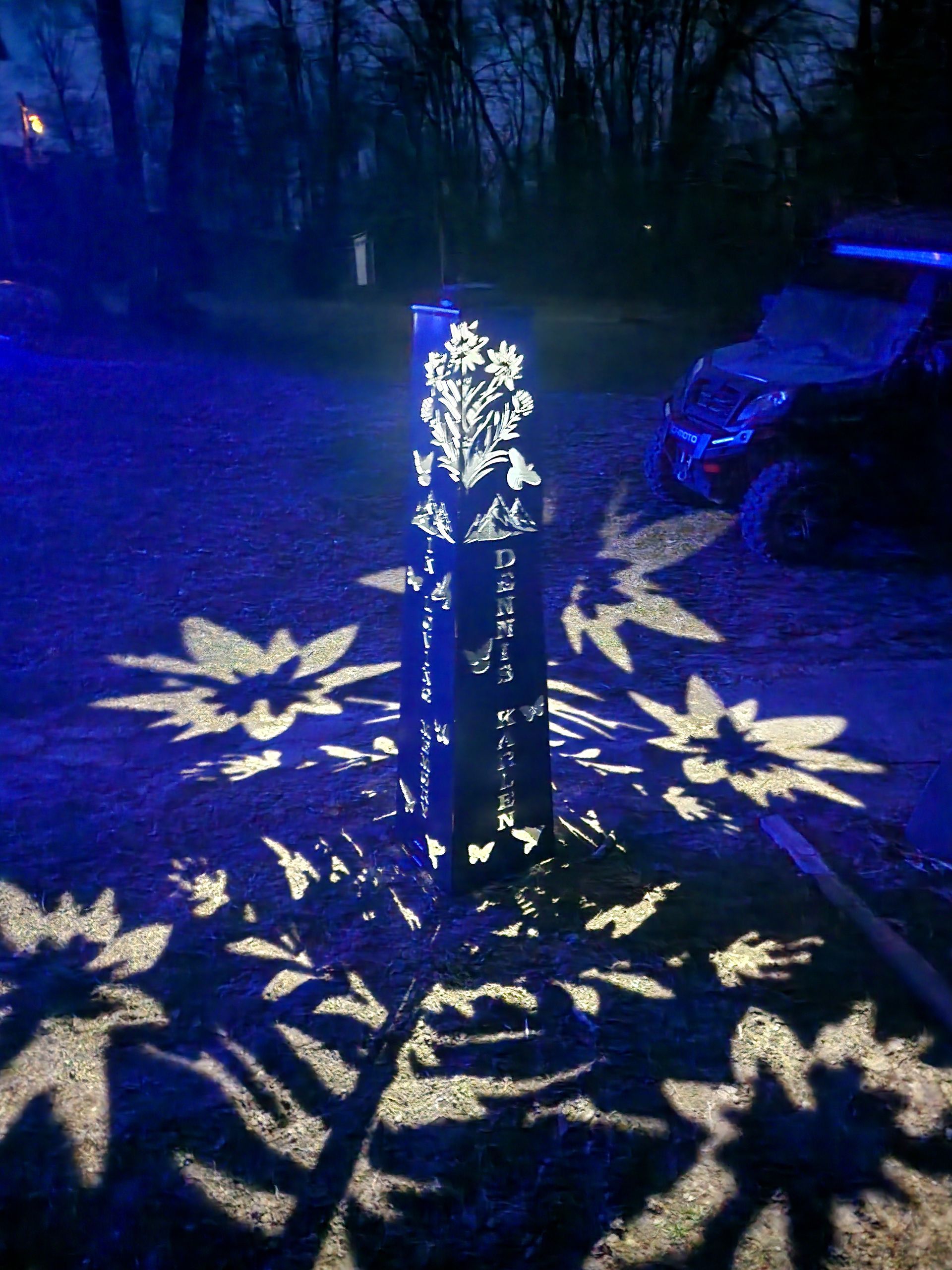 A metal pillar with floral cutouts projects flower-shaped shadows on pavement, lit by blue light at night.