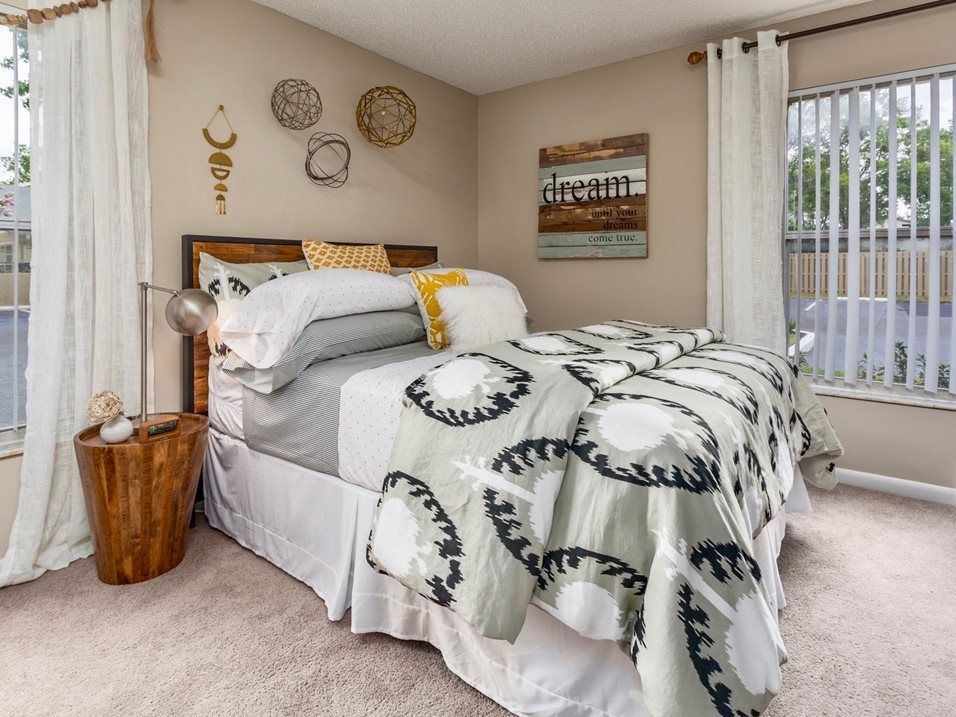 Bedroom with bed, patterned comforter, side table, window with blinds, and decorative wall hangings.