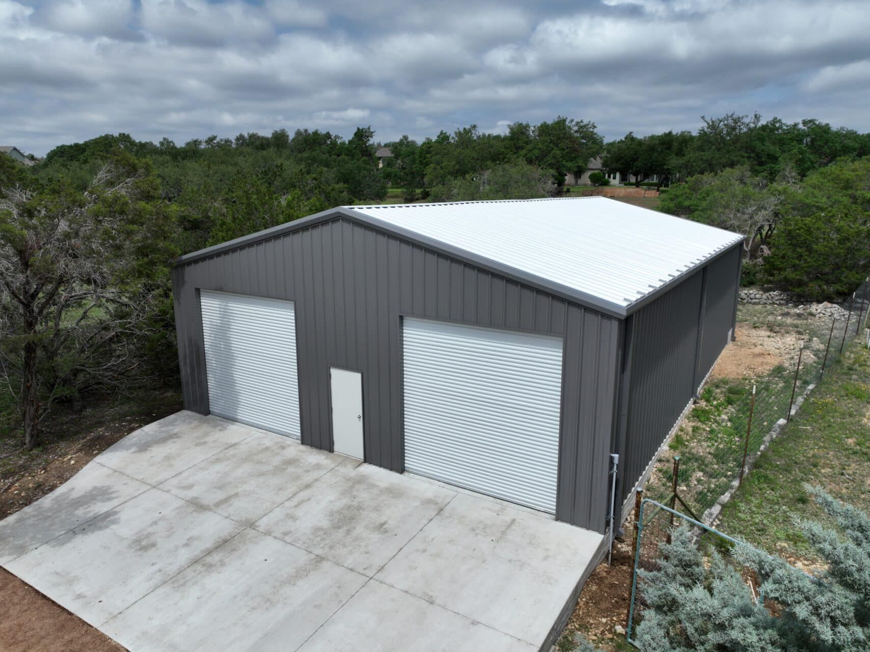 Gray metal building with two garage doors and concrete driveway. Trees in background.