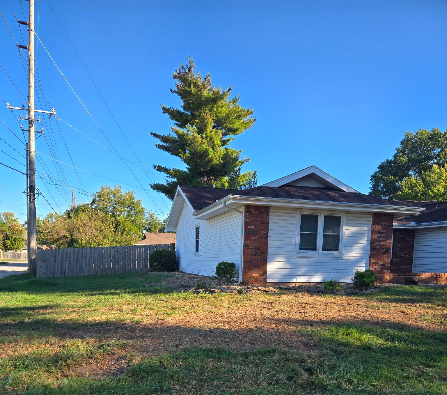 A one-story house with white siding and a brick facade sits under a blue sky, next to a wooden fence.