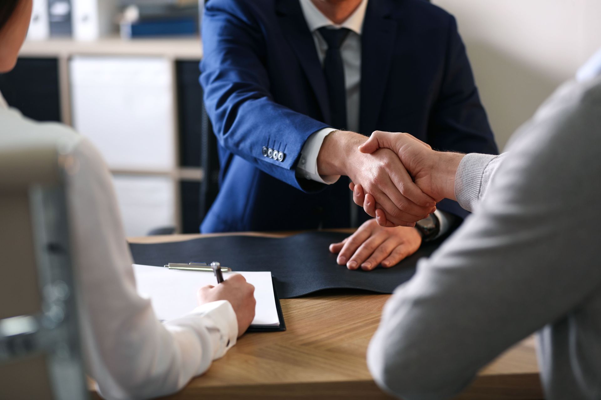 Three people in a meeting, two men shaking hands over a desk, a woman writing.
