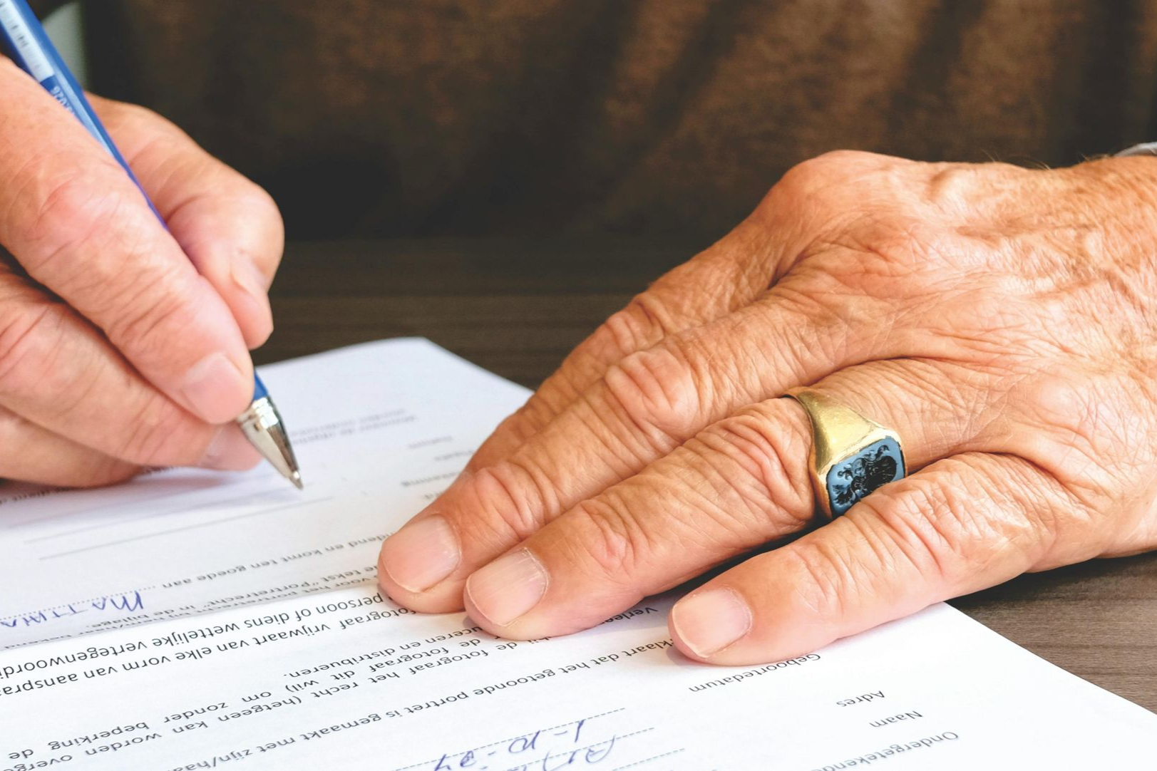 Person's hands signing a document with a pen; they have a ring on one finger, and the paper is on a dark surface.
