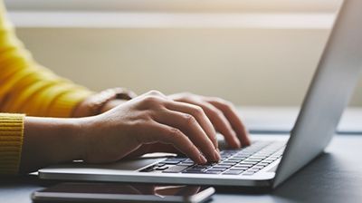 Hands typing on a laptop beside a smartphone on a desk, with a yellow sweater sleeve visible.