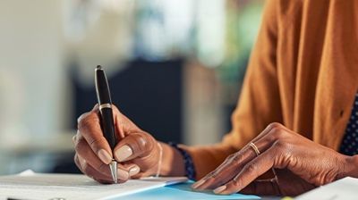 Person writing with a pen on paper at a desk, wearing an orange top.