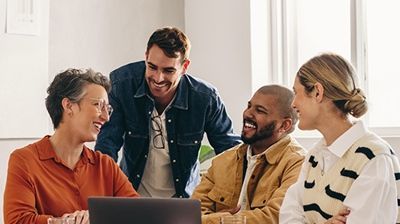 Four coworkers smiling and talking around a laptop in a bright office