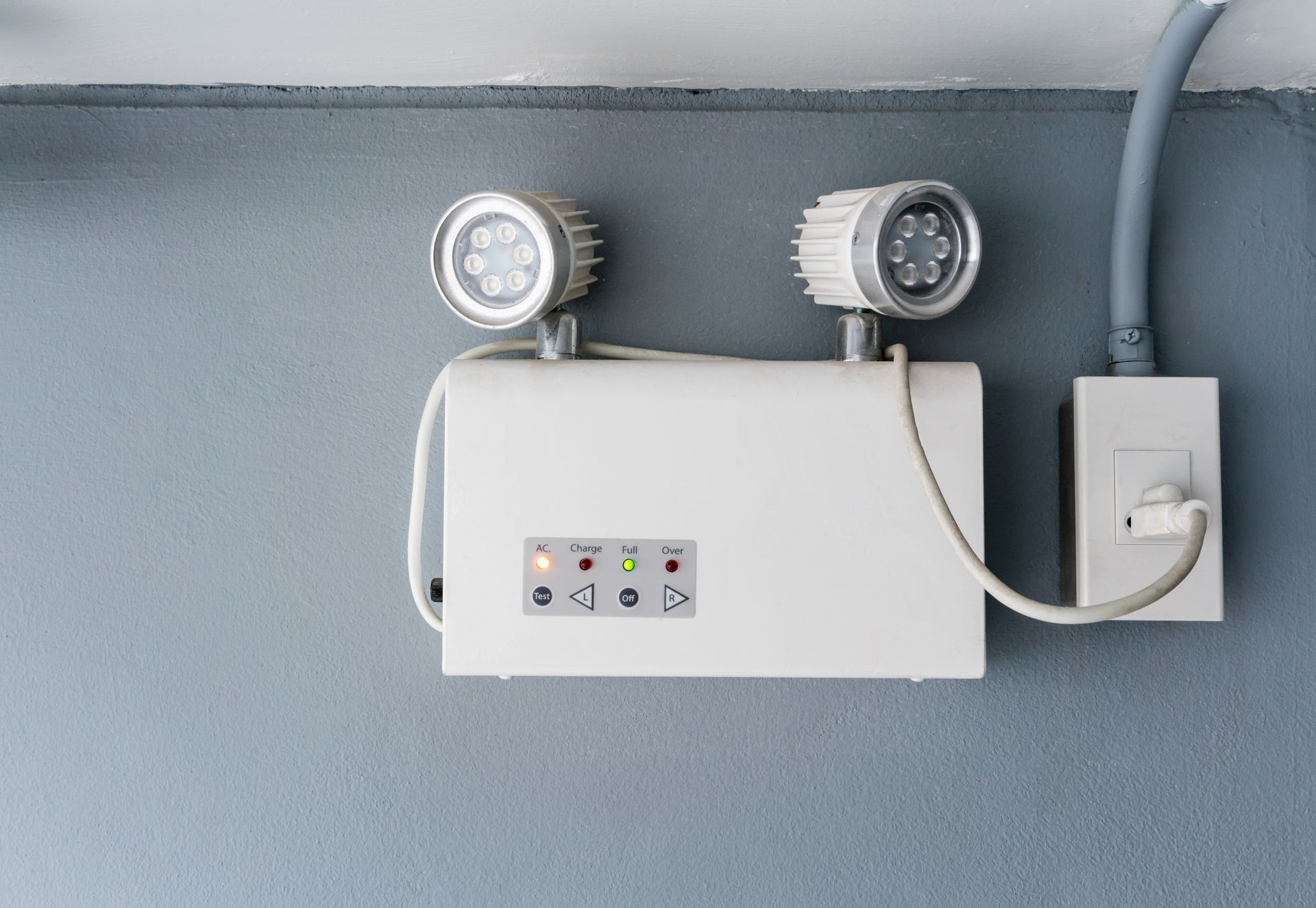 Emergency exit light on a gray wall, white fixture with two lights and a power outlet.