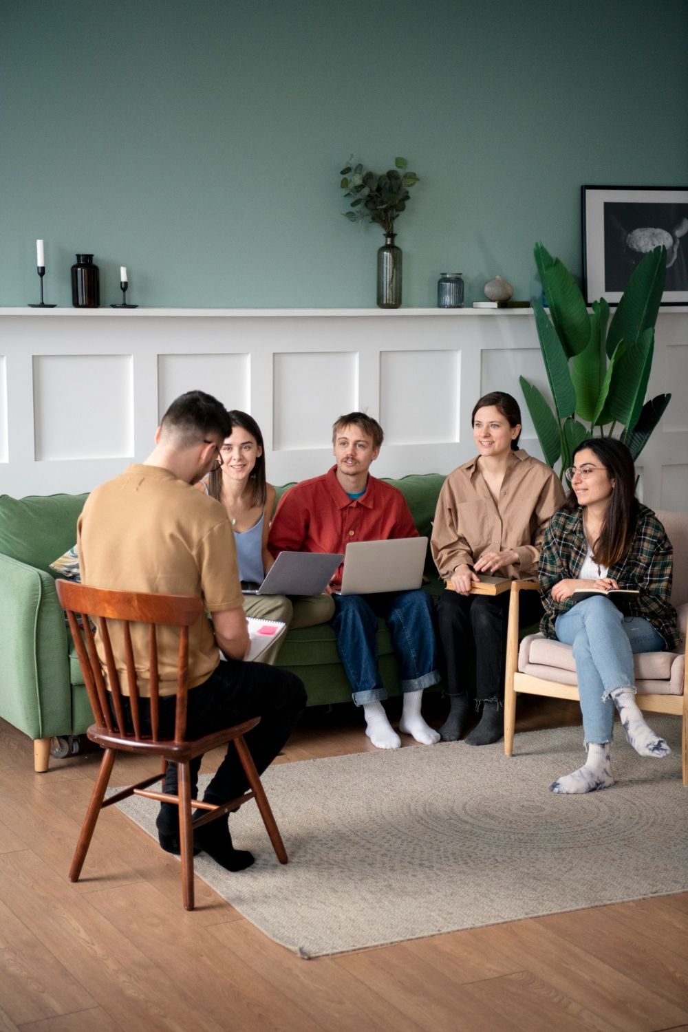 Group meeting in a living room, with four people seated on a couch and one facing them in a chair with a laptop.
