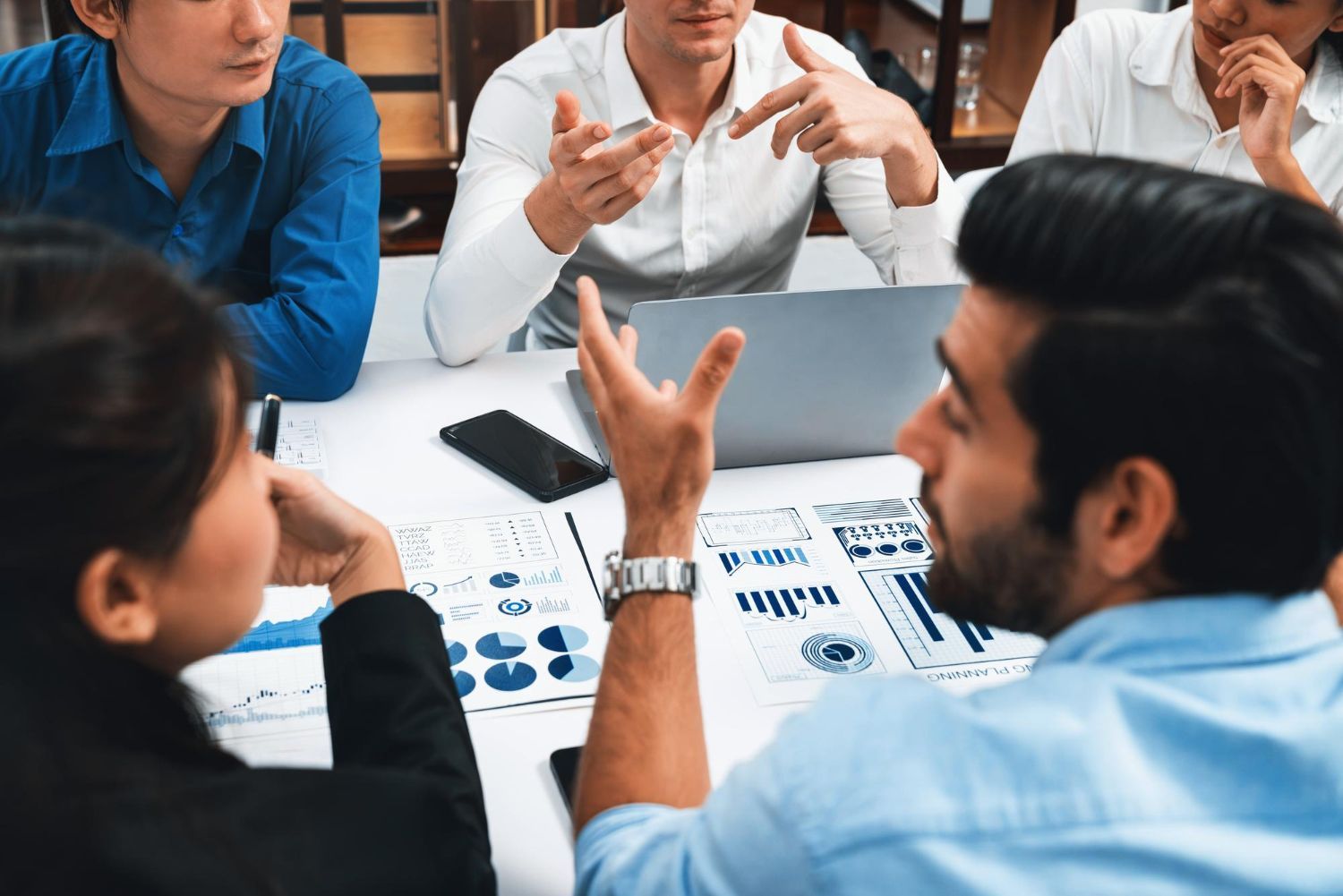 Small group discussing charts around a table in a meeting room, with one person gesturing during the conversation