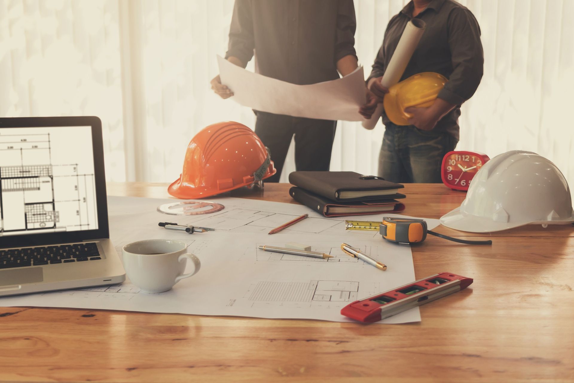 Construction plans on a desk with hard hats, tools, laptop, and two people reviewing papers